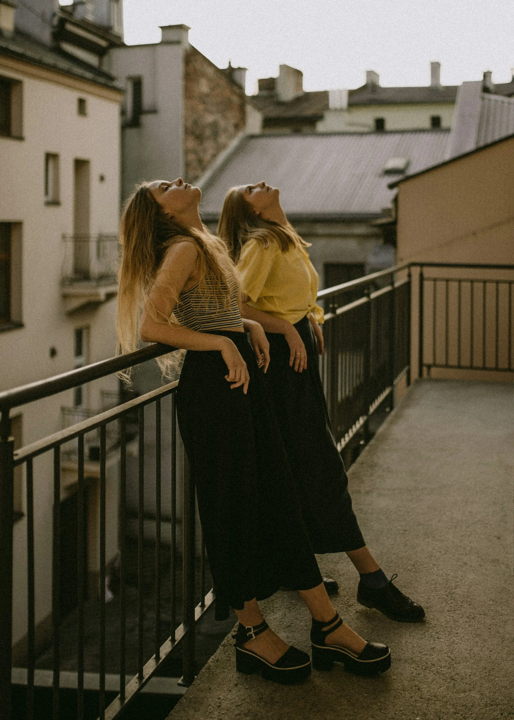 Two women standing on a rooftop balcony, leaning against the railing with their heads tilted back and eyes closed, enjoying the sunset or evening light in an urban setting with apartment buildings in the background.