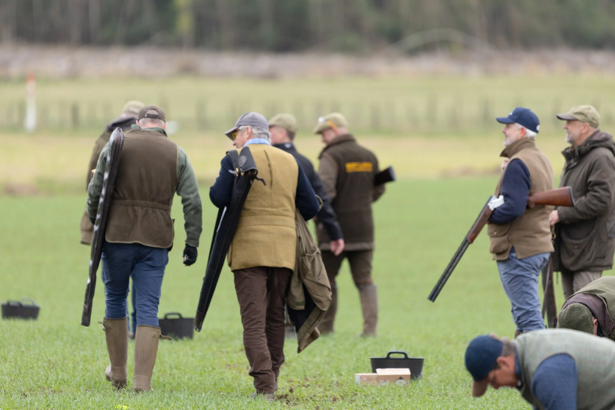 Group of men in outdoor clothing and hats standing on a grassy field, some holding rifles, participating in a bird hunting or shooting activity, with a body of water and trees in the background.