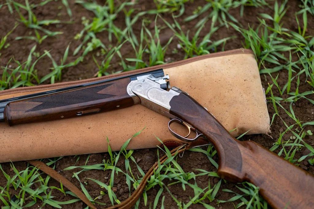 A shotgun resting on a tan gun case on the ground with green grass and soil.