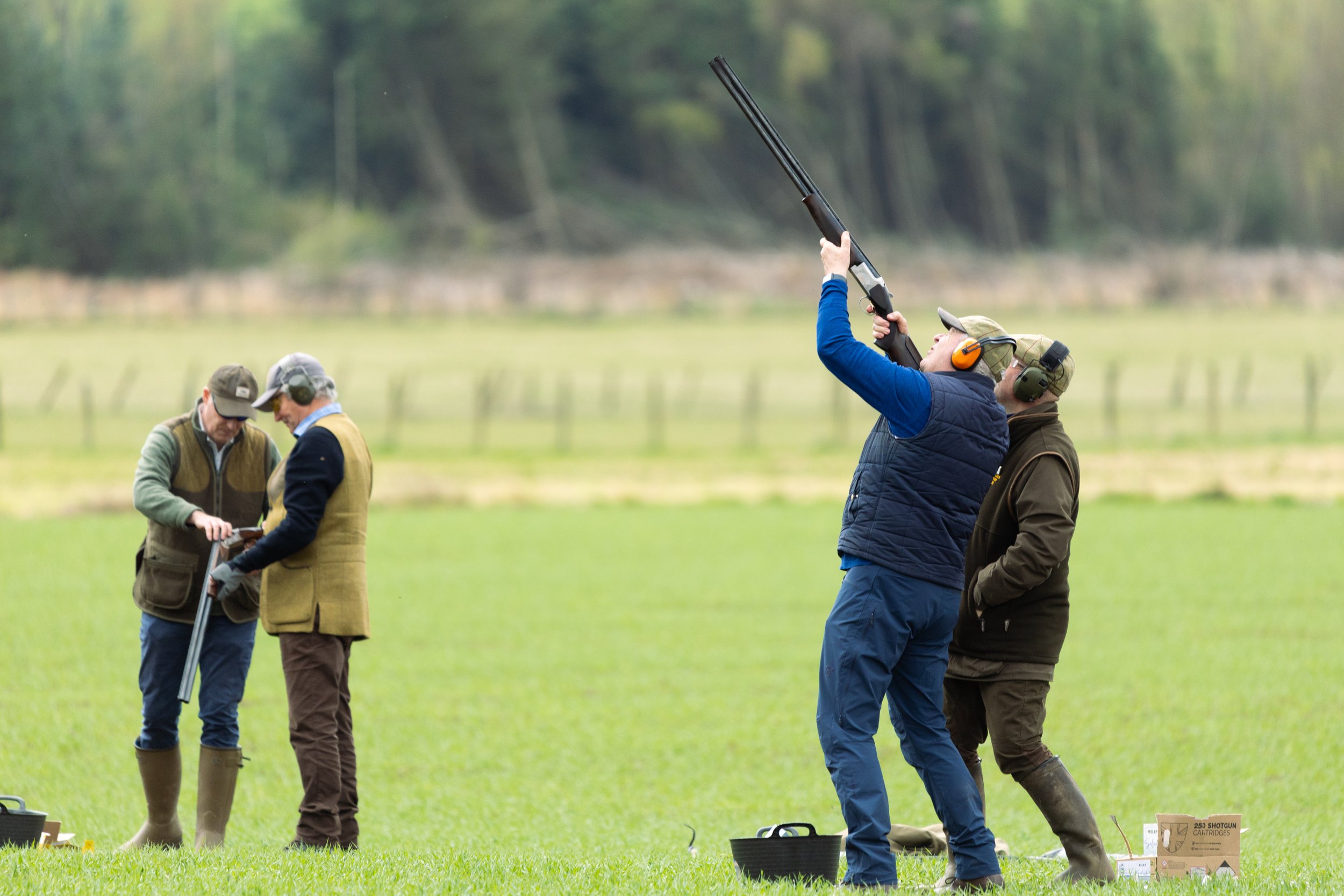 People shooting shotguns at a rural outdoor range, wearing ear protection and jackets, with boxes of ammunition nearby, and two other individuals standing and discussing equipment in the background.