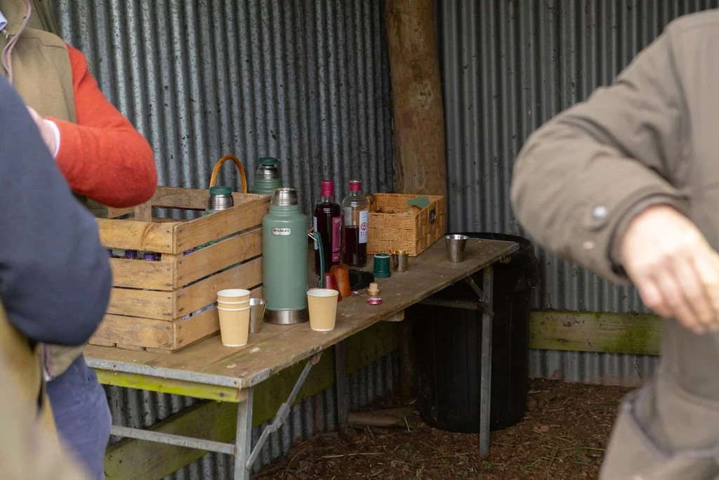 A rustic wooden table with thermoses, bottles, paper cups, and small containers inside a corrugated metal shed.