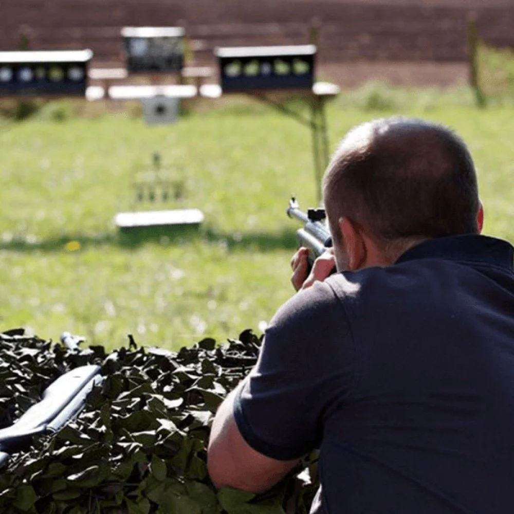 A man aiming a rifle at a distant target during shooting practice outdoors. There are benches and targets in the background, with the man lying behind a hedge.