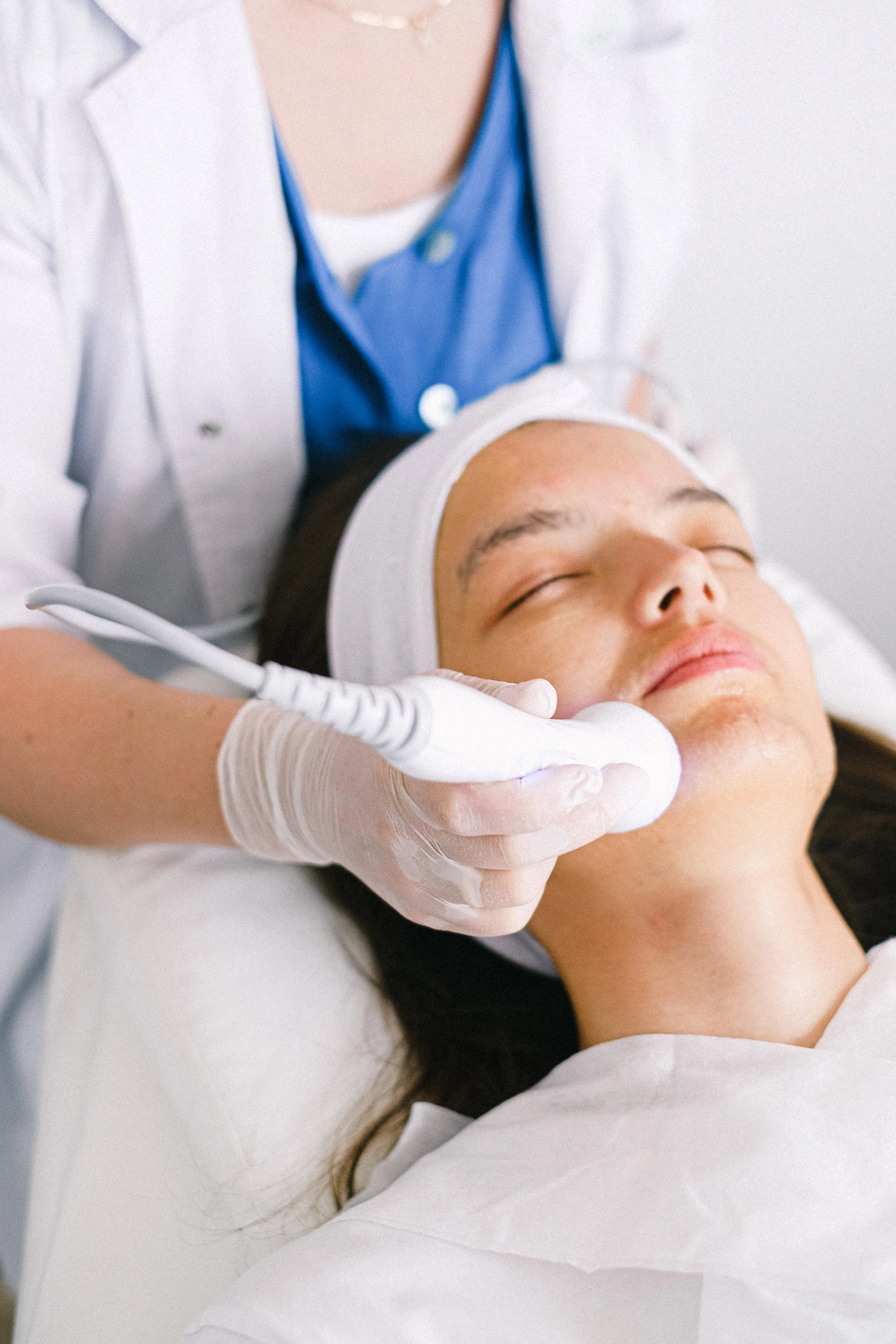 A woman receiving a facial treatment in a spa or clinic, lying down with eyes closed, while a technician applies a facial device.