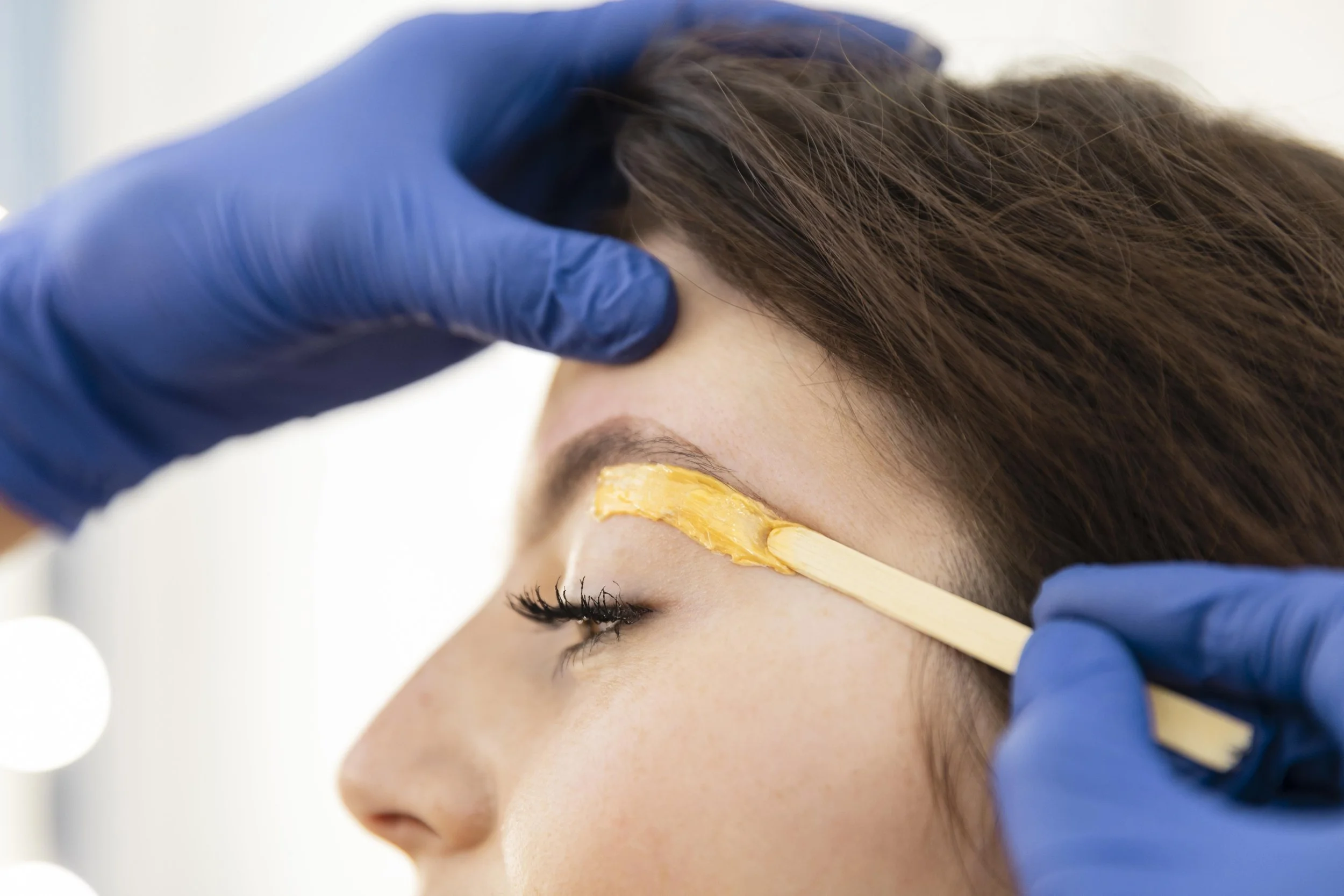 A person getting beauty treatment on their eyebrows with a spatula applying a wax or similar substance while another hand holds their forehead, with all hands wearing blue gloves.