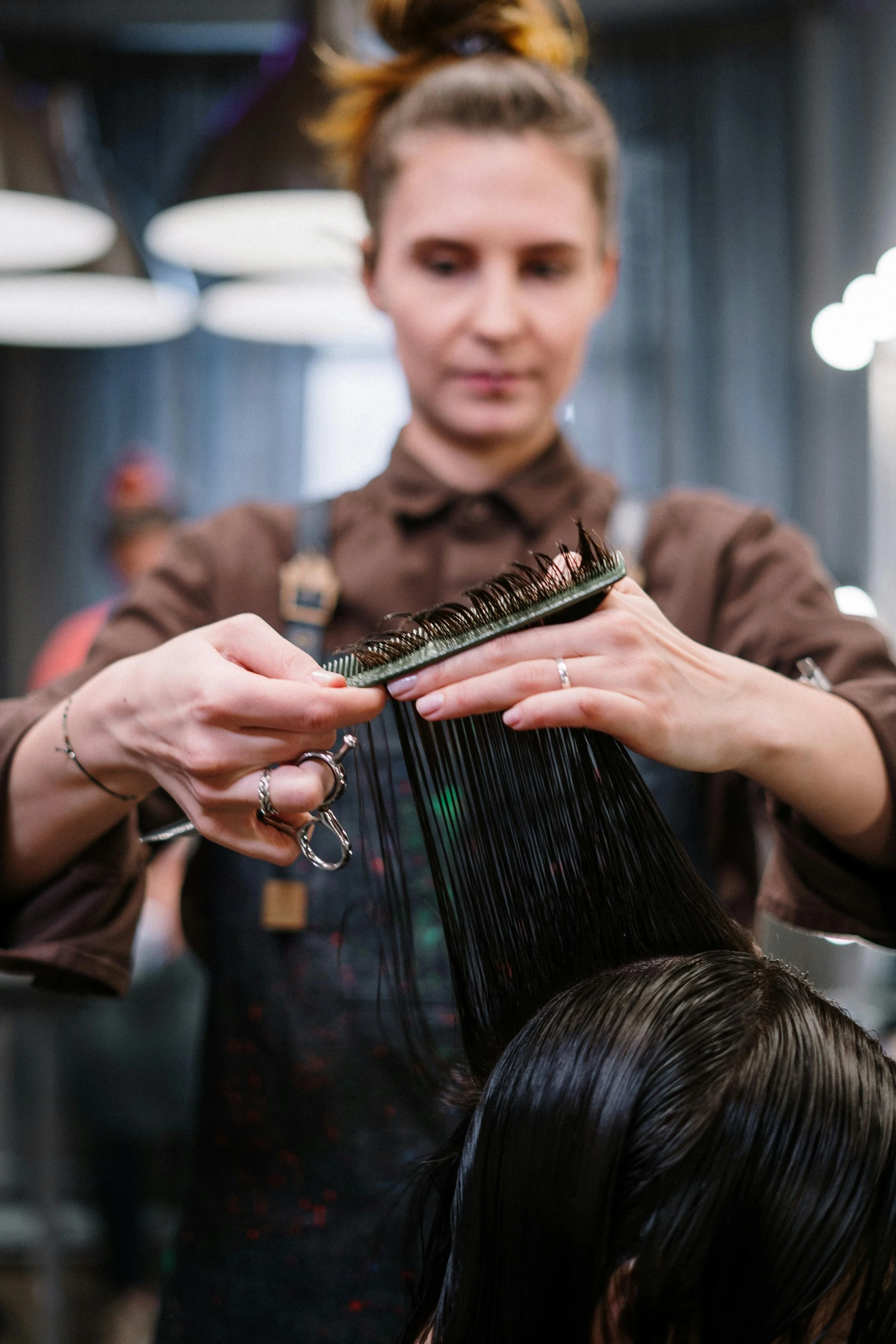 A hairdresser combs a customer's dark, wet hair in a salon, with salon lighting in the background.