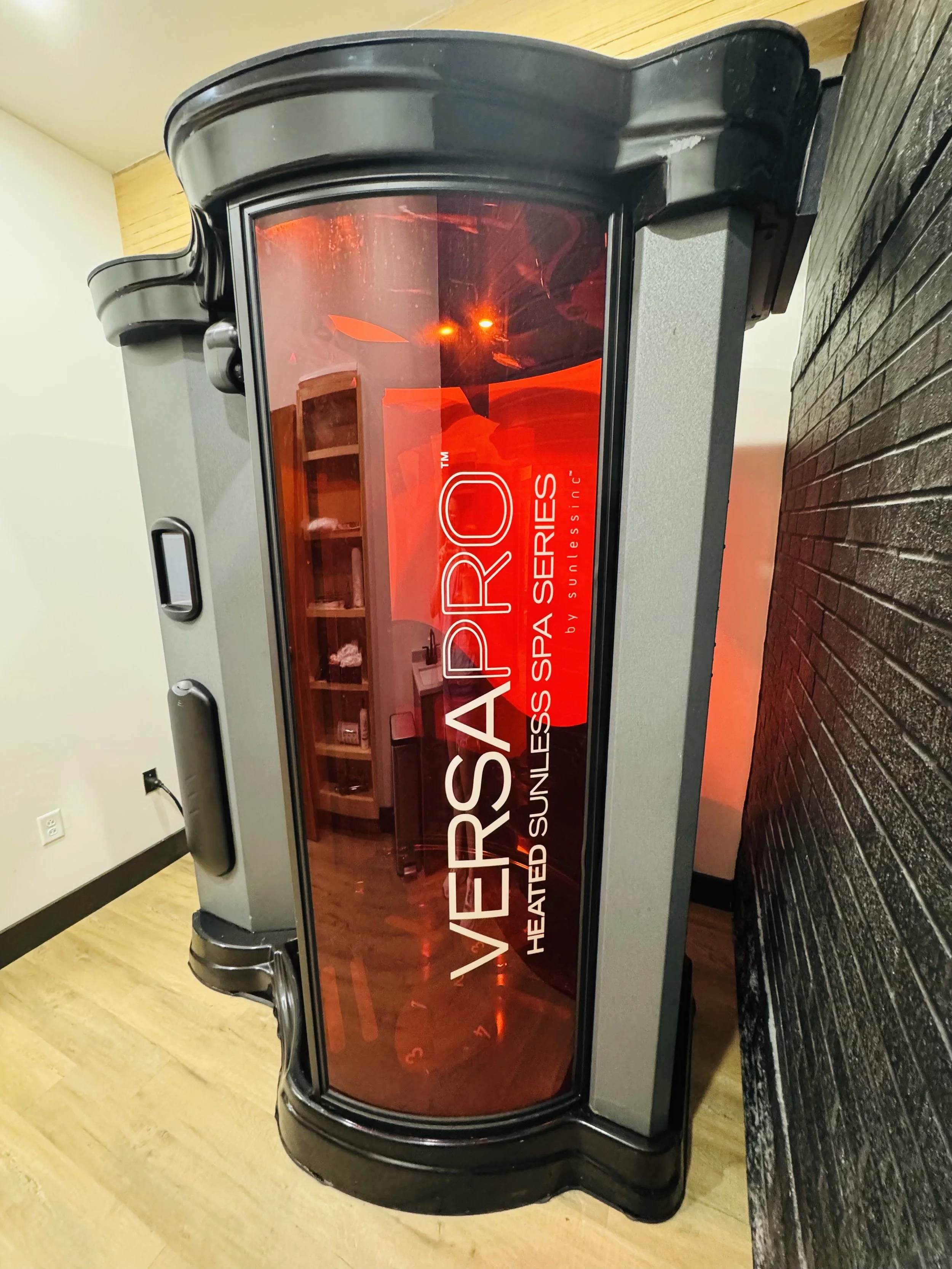 Heated sunless spray tanning machine labeled VersaPro by Sunless Series in a room with a wooden floor and a black brick wall.