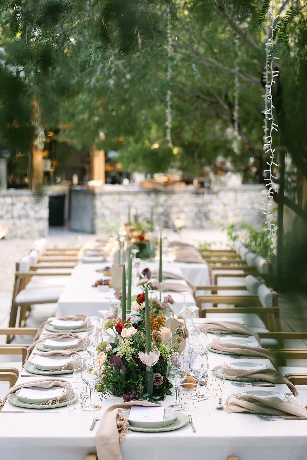 Outdoor dining table set for a celebration, decorated with flower arrangements and candles, surrounded by chairs with beige napkins, under trees with hanging string lights.