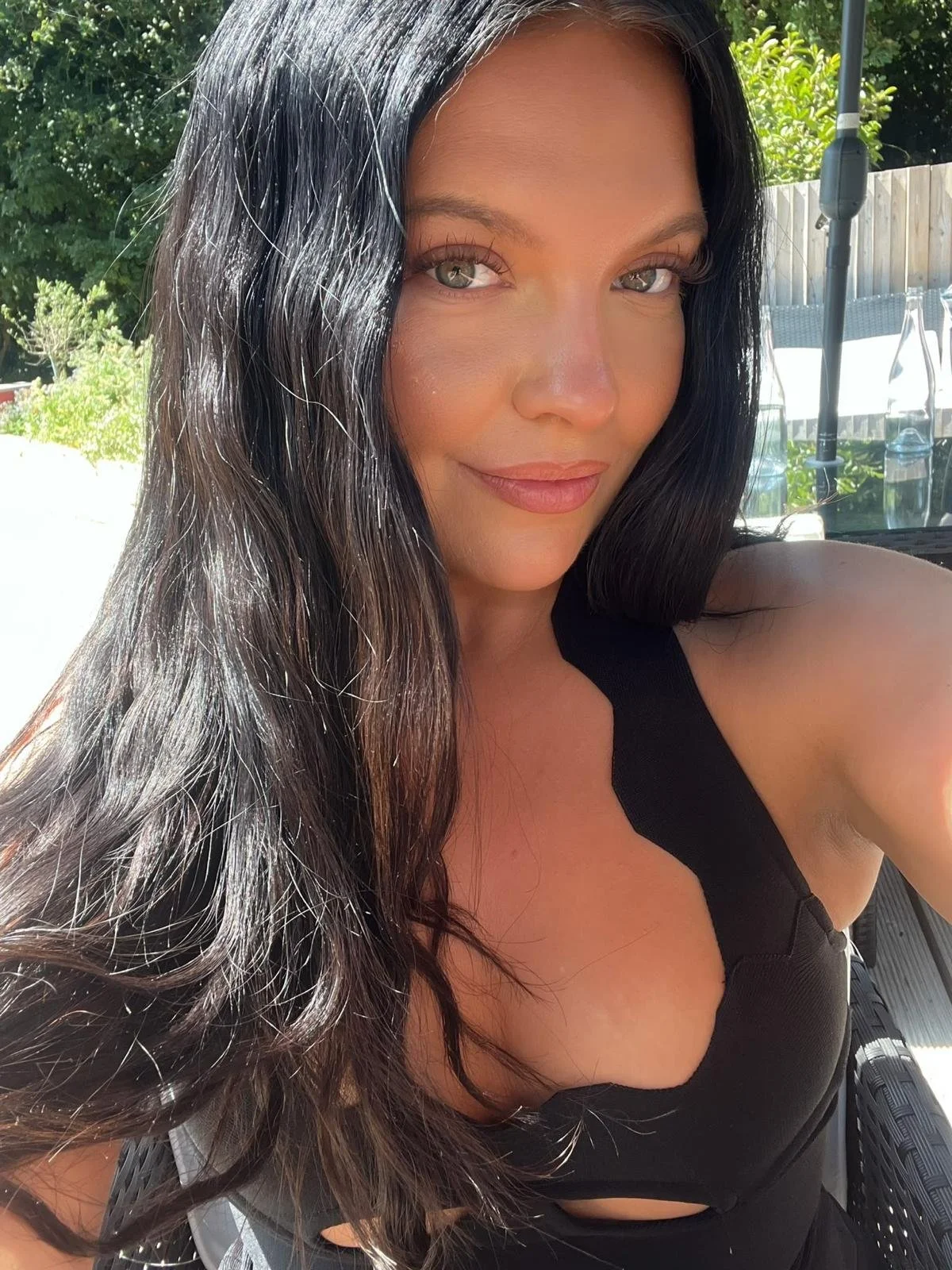 A woman with long dark hair and light skin, smiling outdoors in sunlight, wearing a black top, with greenery and outdoor furniture in the background.