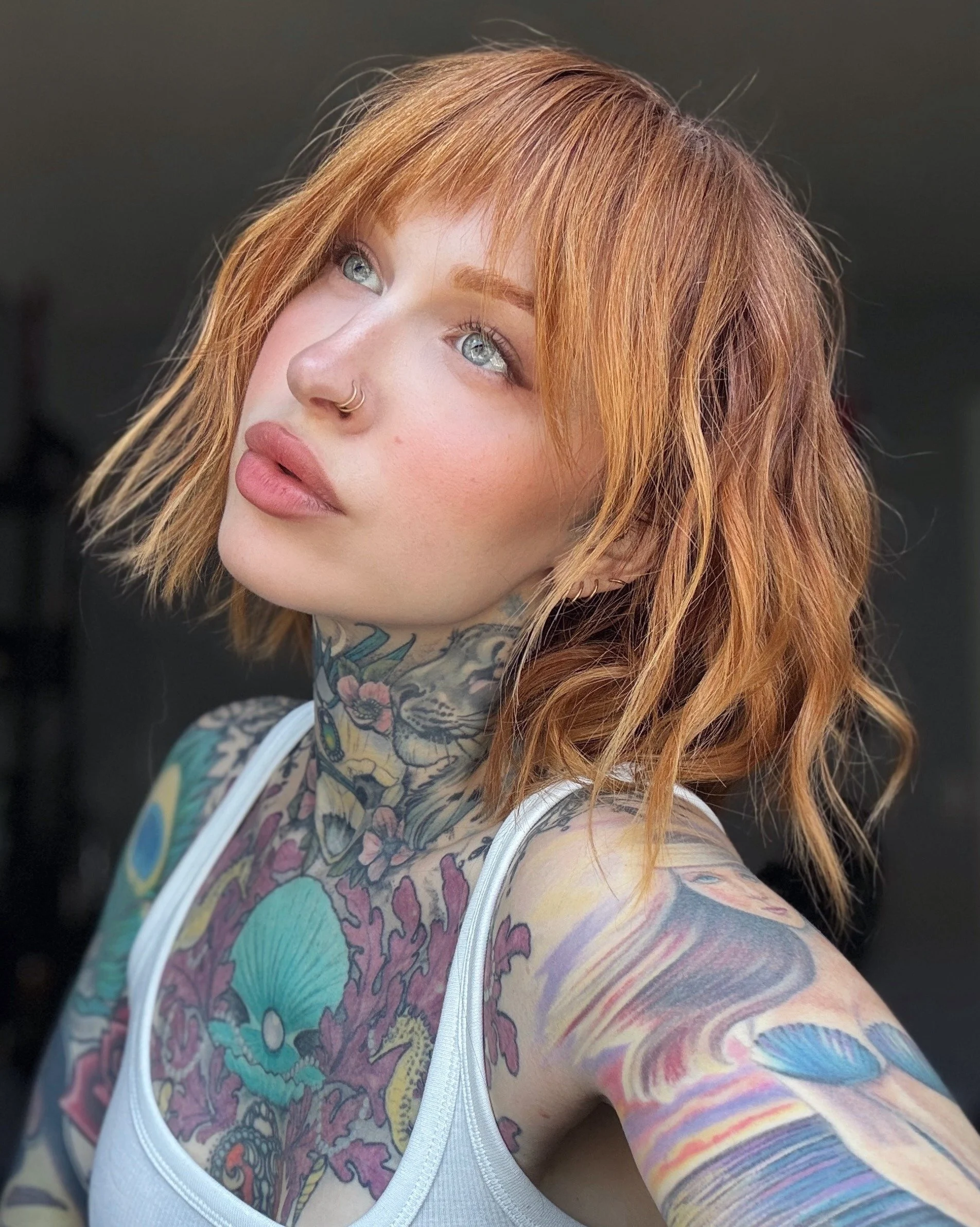 A woman with red hair and tattoos on her neck and arms, wearing a white tank top, looks upwards and to the left. The background is dark and out of focus.