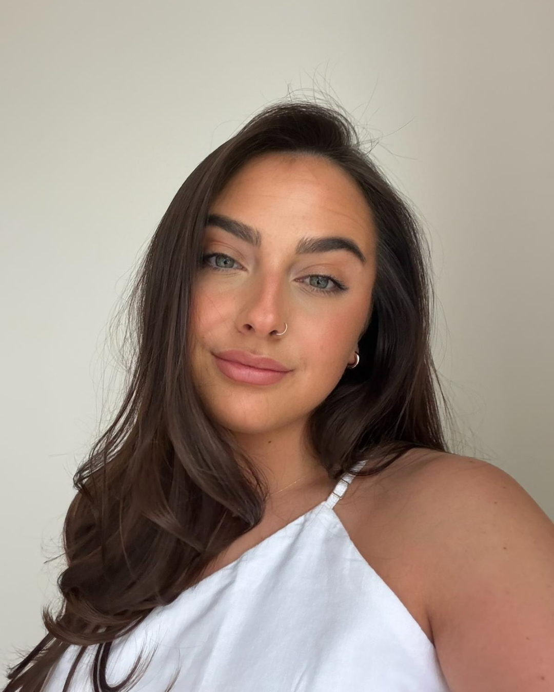 A young woman with long, wavy dark brown hair and piercing blue eyes, wearing a white top with one shoulder visible, standing against a plain light-colored background.