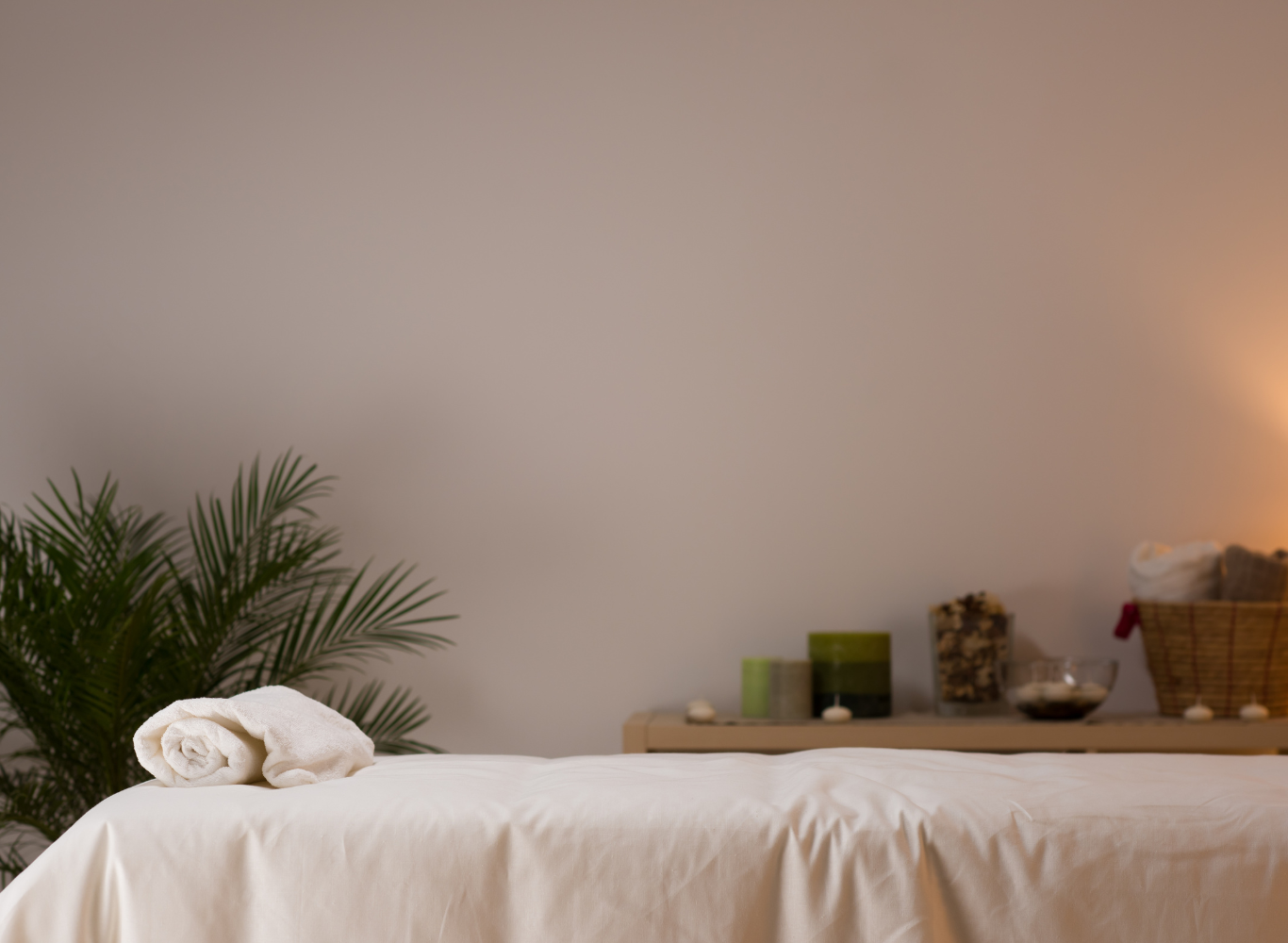 A massage table with a rolled white towel, a green potted plant on the left, and a wooden shelf with candles, a basket of towels, and bowls on the right. The room has soft lighting and a neutral wall.