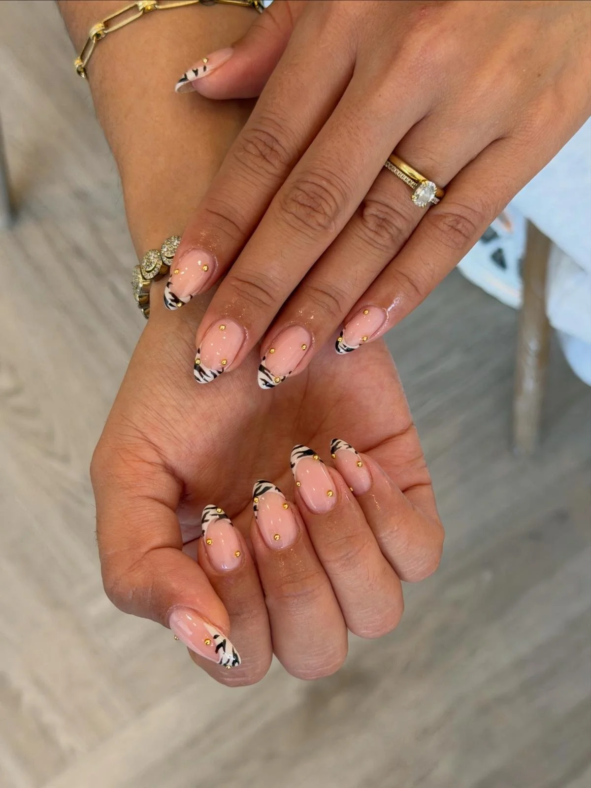Close-up of two hands with manicured nails featuring zebra-striped tips and small yellow dots, adorned with rings and jewelry. One hand is resting on top of the other, displaying the nail art and rings.