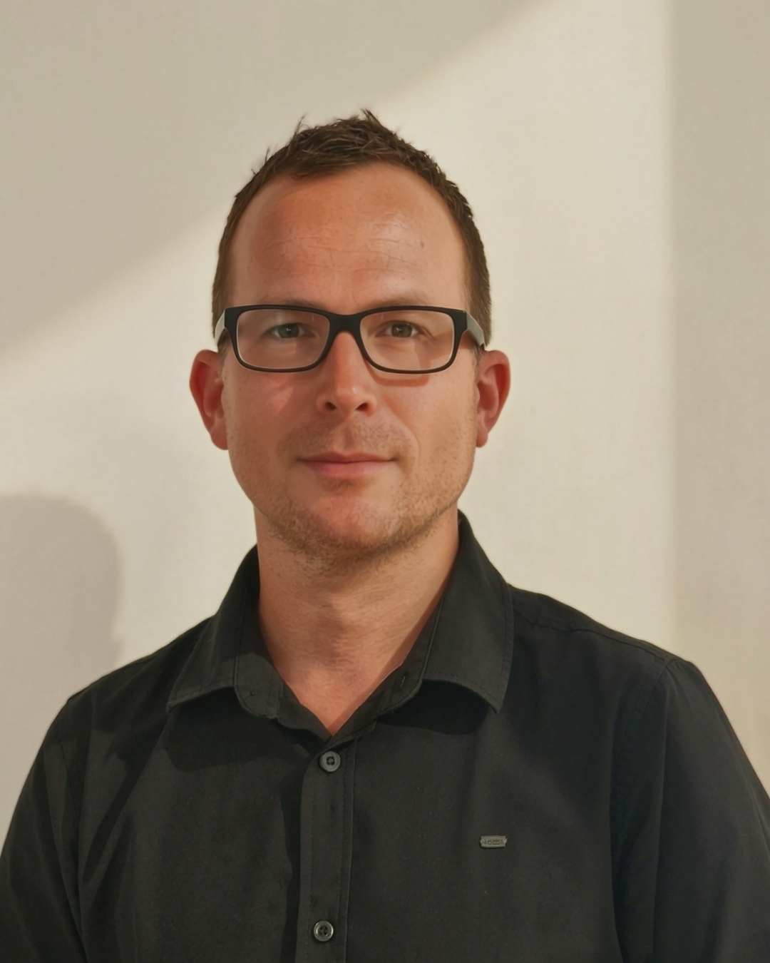 Portrait of a man with short brown hair, glasses, and a slight smile, wearing a black button-up shirt against a plain background.