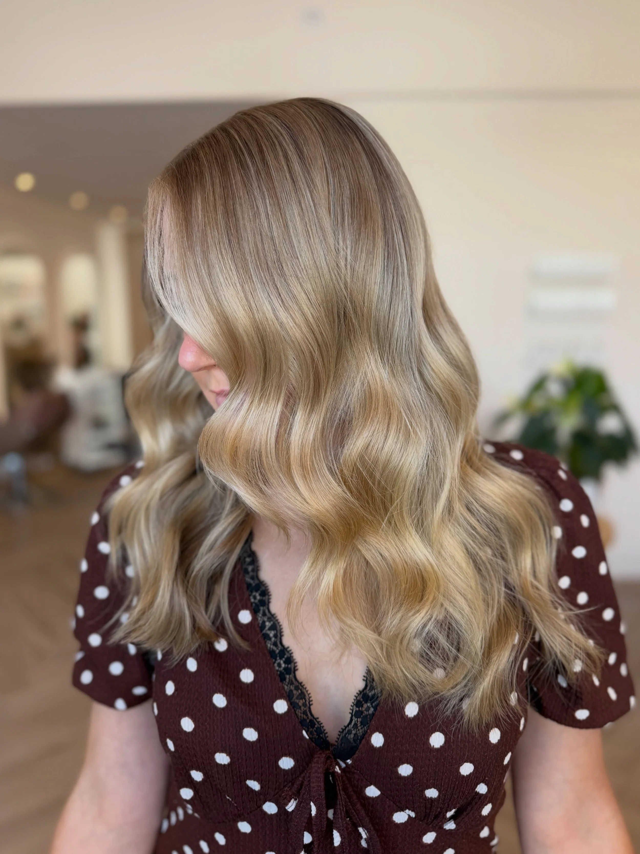 Woman with wavy blonde hair wearing a brown dress with white polka dots and lace detailing.