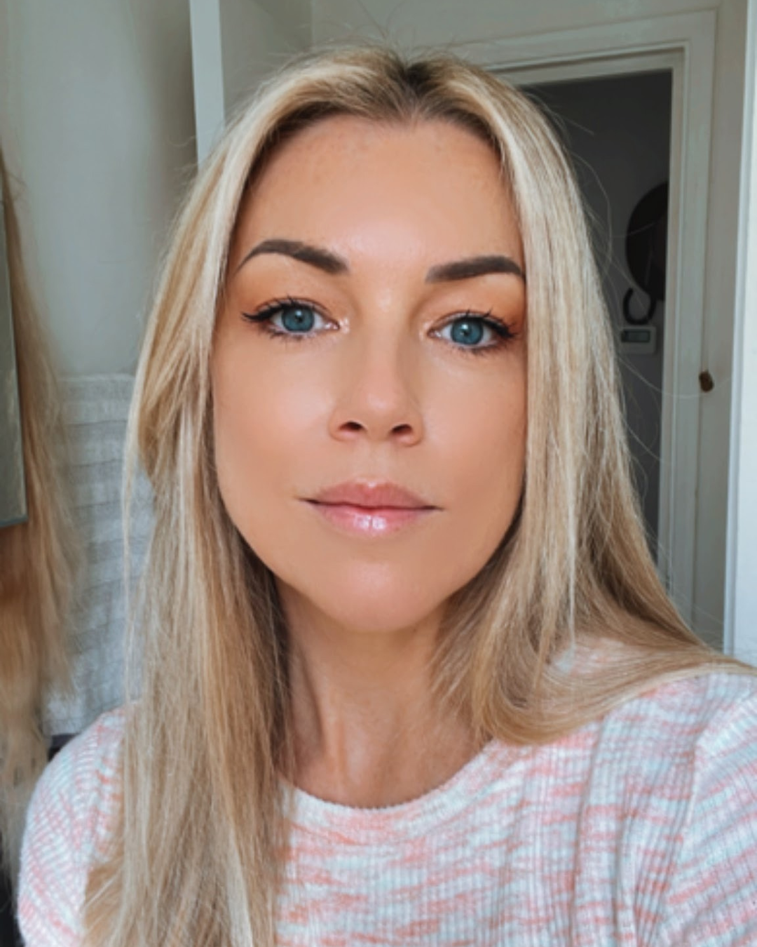 A close-up of a woman with long blonde hair, blue eyes, light makeup, wearing a pink and white checkered shirt, indoors with a light-colored wall and door in the background.