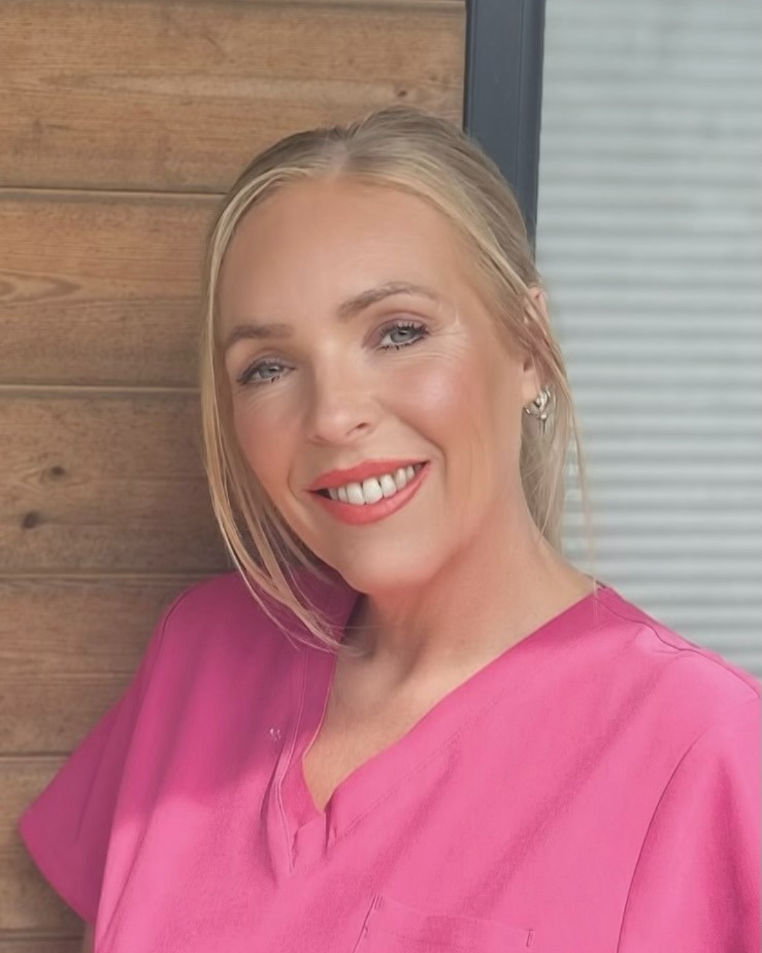 A smiling woman with blonde hair in a pink top, standing against a wooden wall and a window with horizontal blinds.