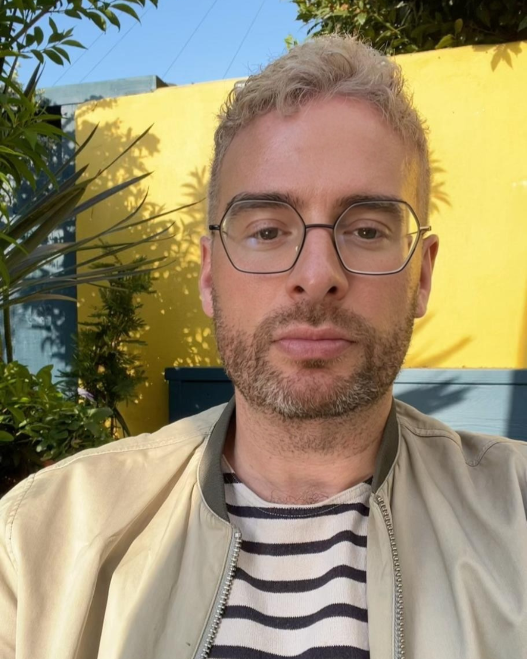 A man with glasses and a beard wearing a beige jacket and striped shirt, sitting outdoors with a yellow wall and plants in the background.