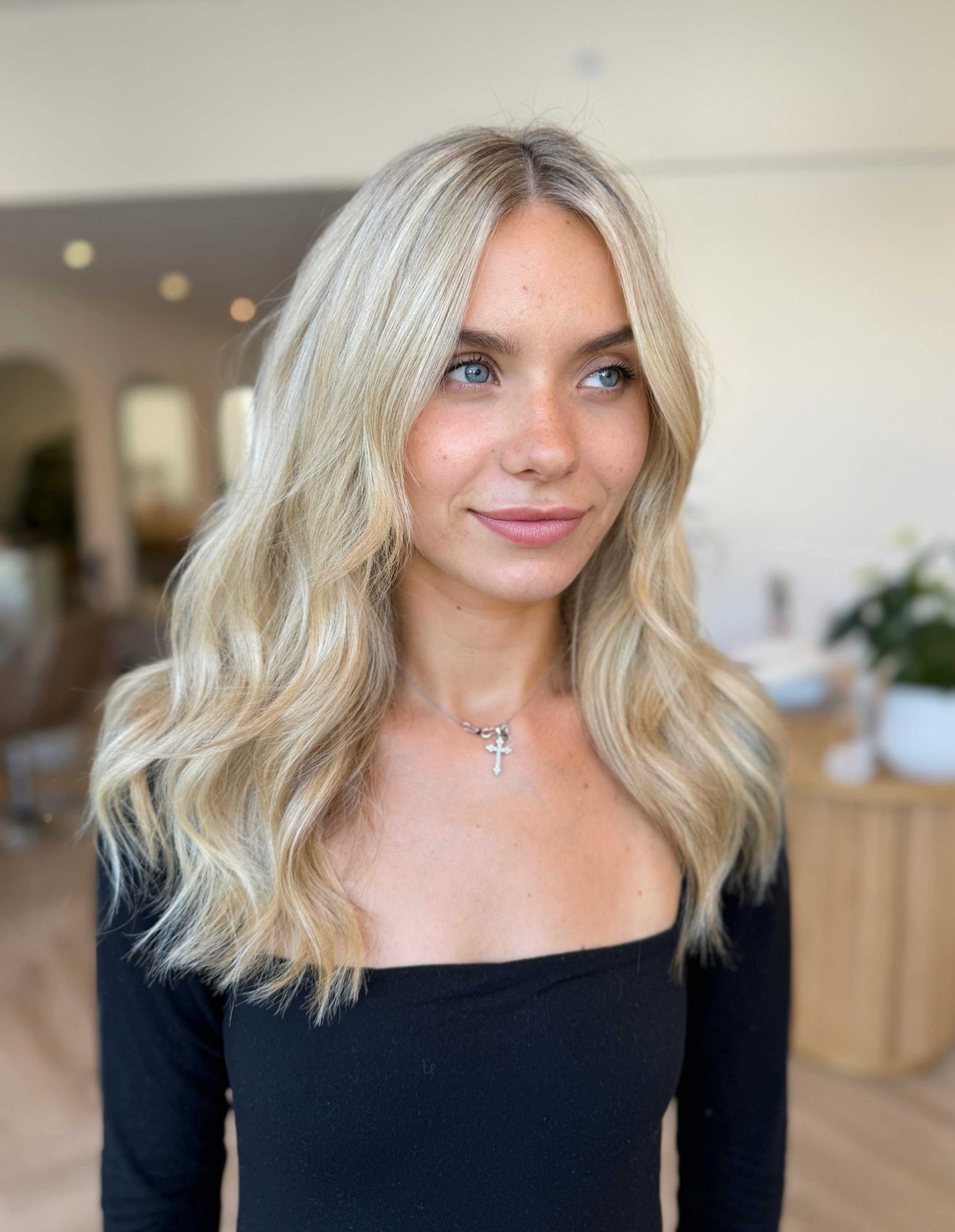 A woman with long, wavy blonde hair, blue eyes, wearing a black top, and a silver cross necklace, standing indoors with a blurred background of a home interior.