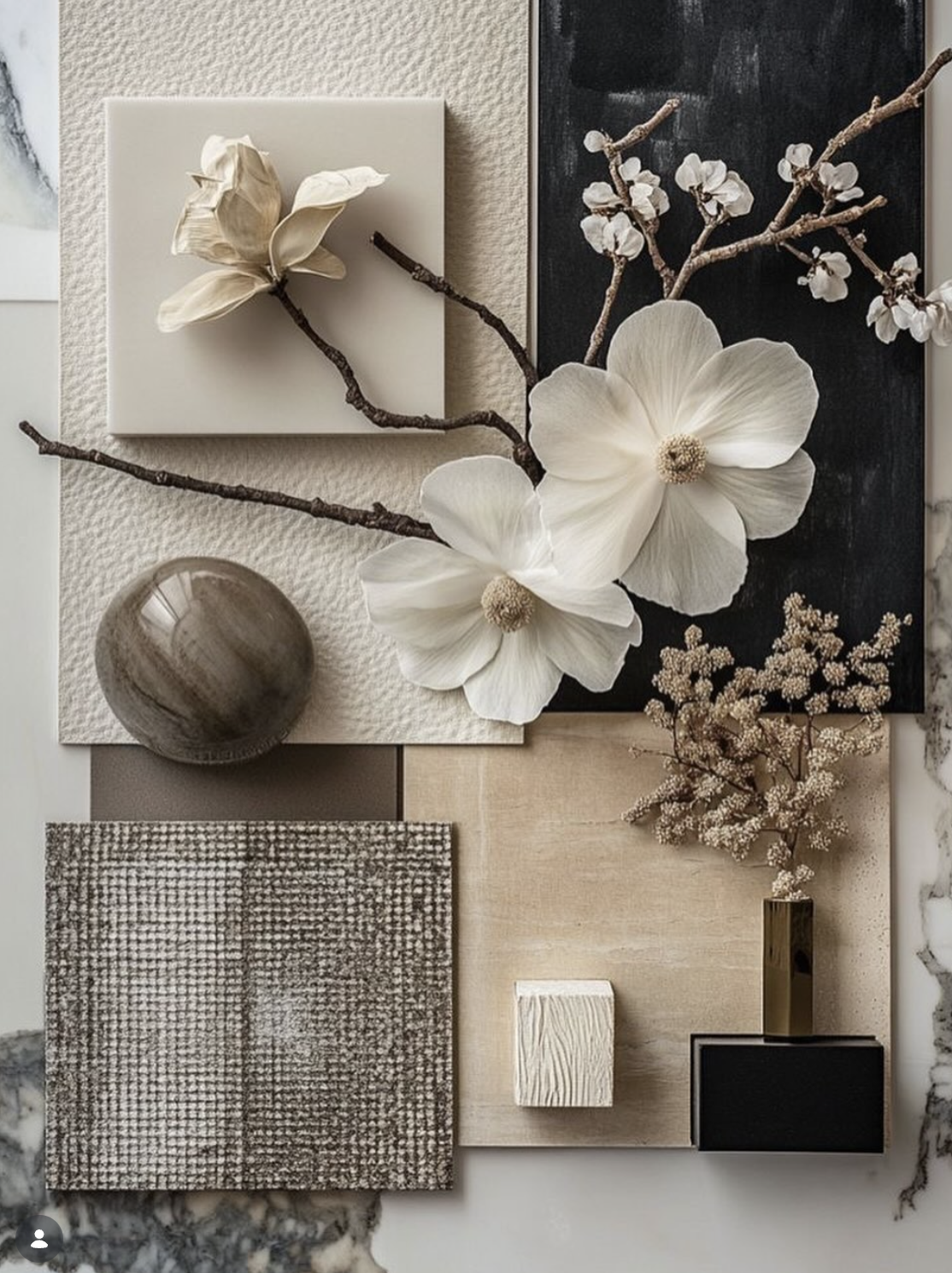 Flat lay of various natural and textured objects, including white magnolia and cherry blossom branches, a black and white textured square, a round marble bowl, a small white textured cube, a wooden tile, a small green vase with cream-colored flowers, and neutral-colored fabric samples on a marble surface.