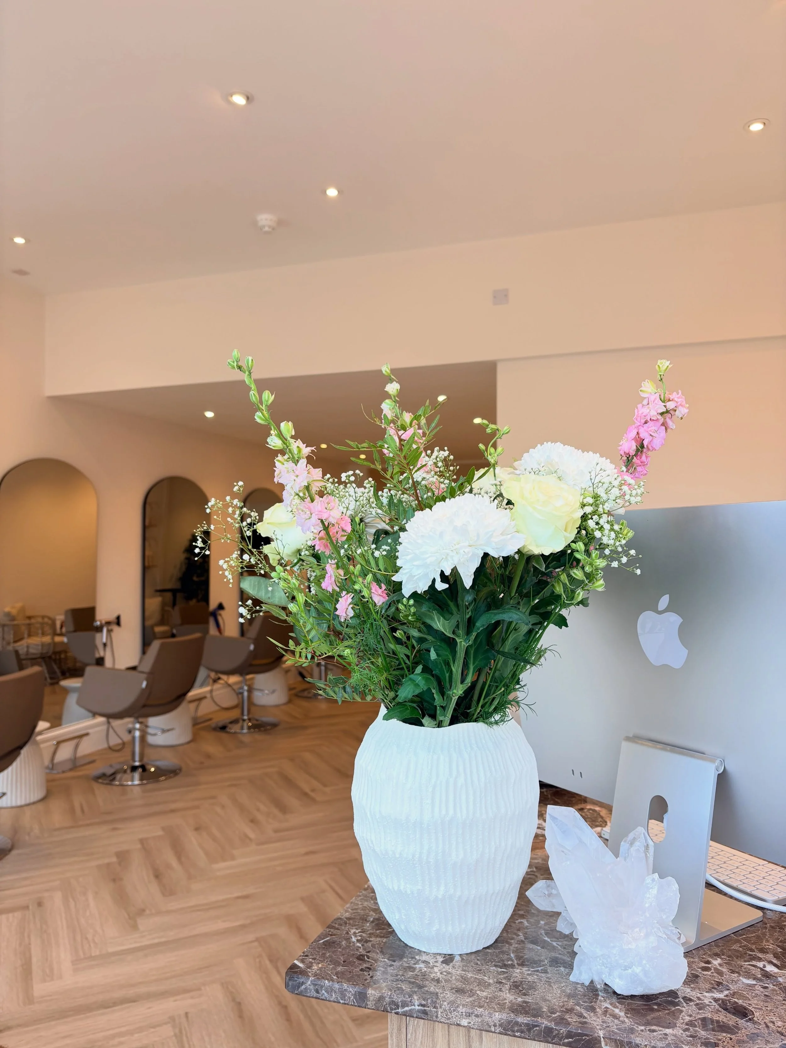 A white textured ceramic vase holding a bouquet of white and pink flowers, including roses and snapdragons, placed on a marble table in a modern, well-lit salon with beige walls and wooden flooring.