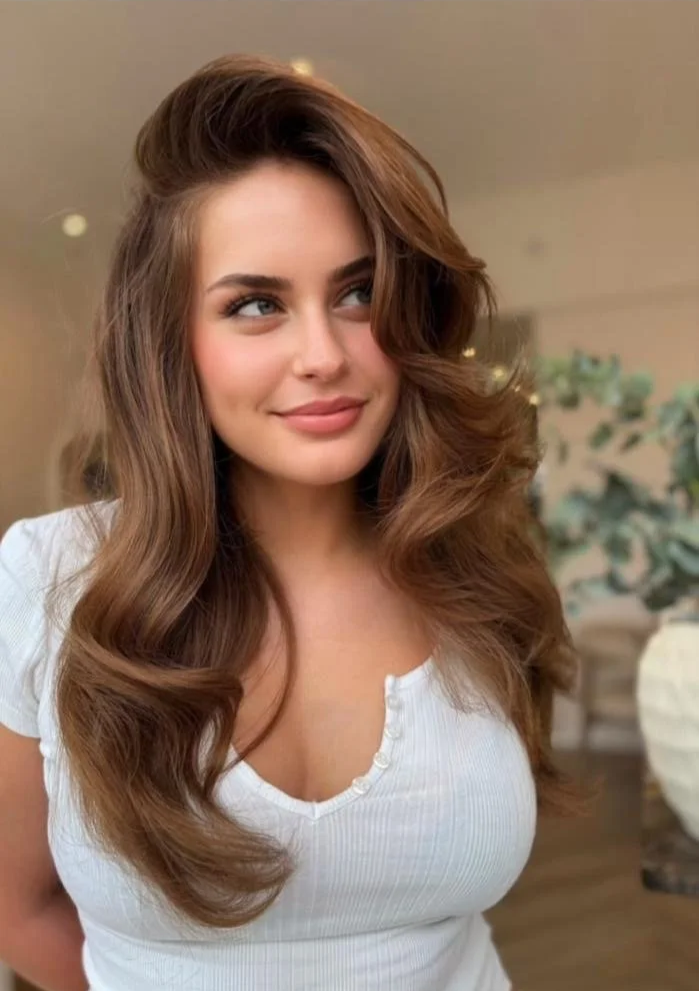 A young woman with long, wavy brown hair and a slight smile, wearing a white top, standing indoors with a blurred background.