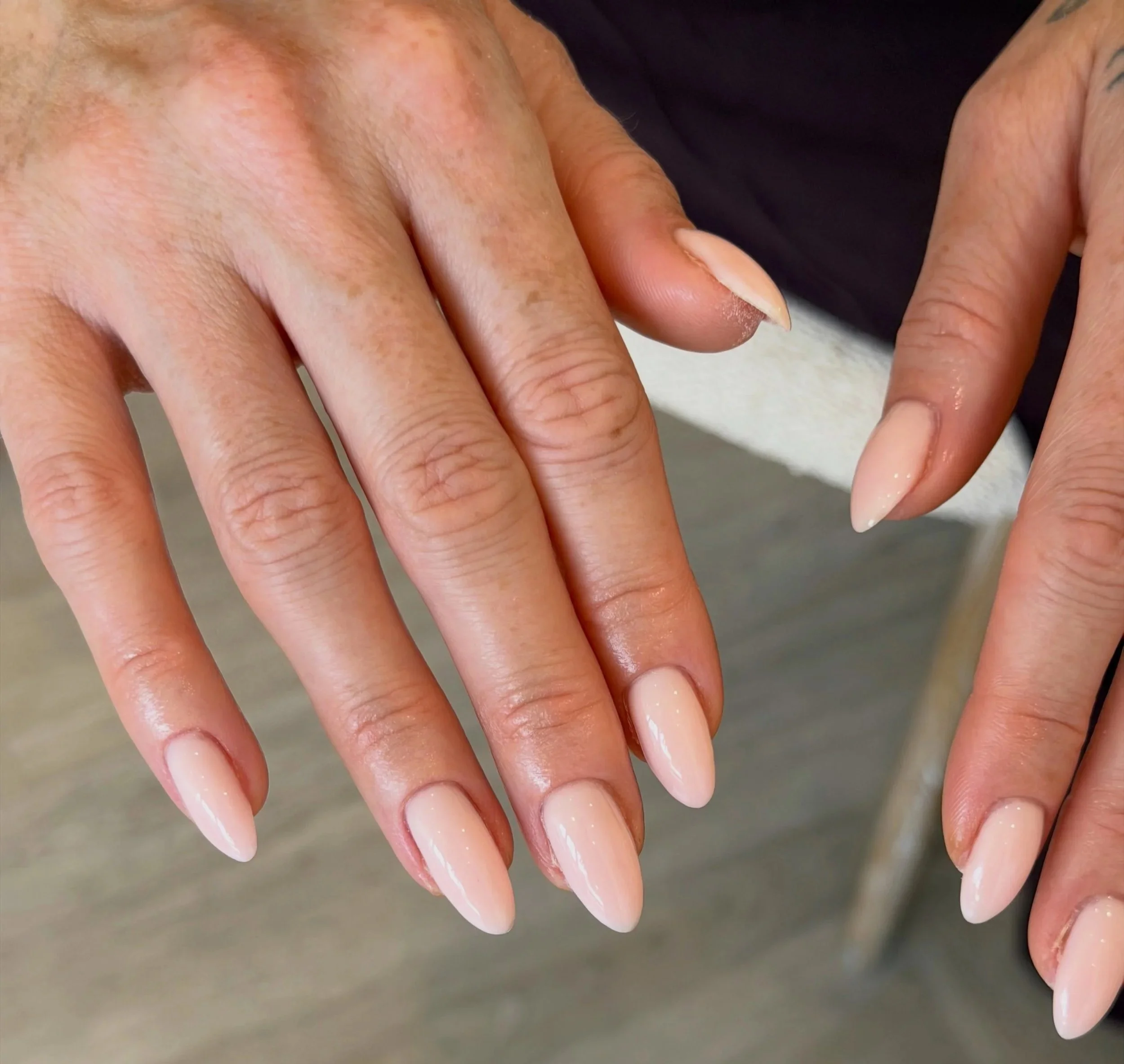 Close-up of two hands with well-manicured, glossy, light pink nails. The skin tone is tan, and the background is blurred.