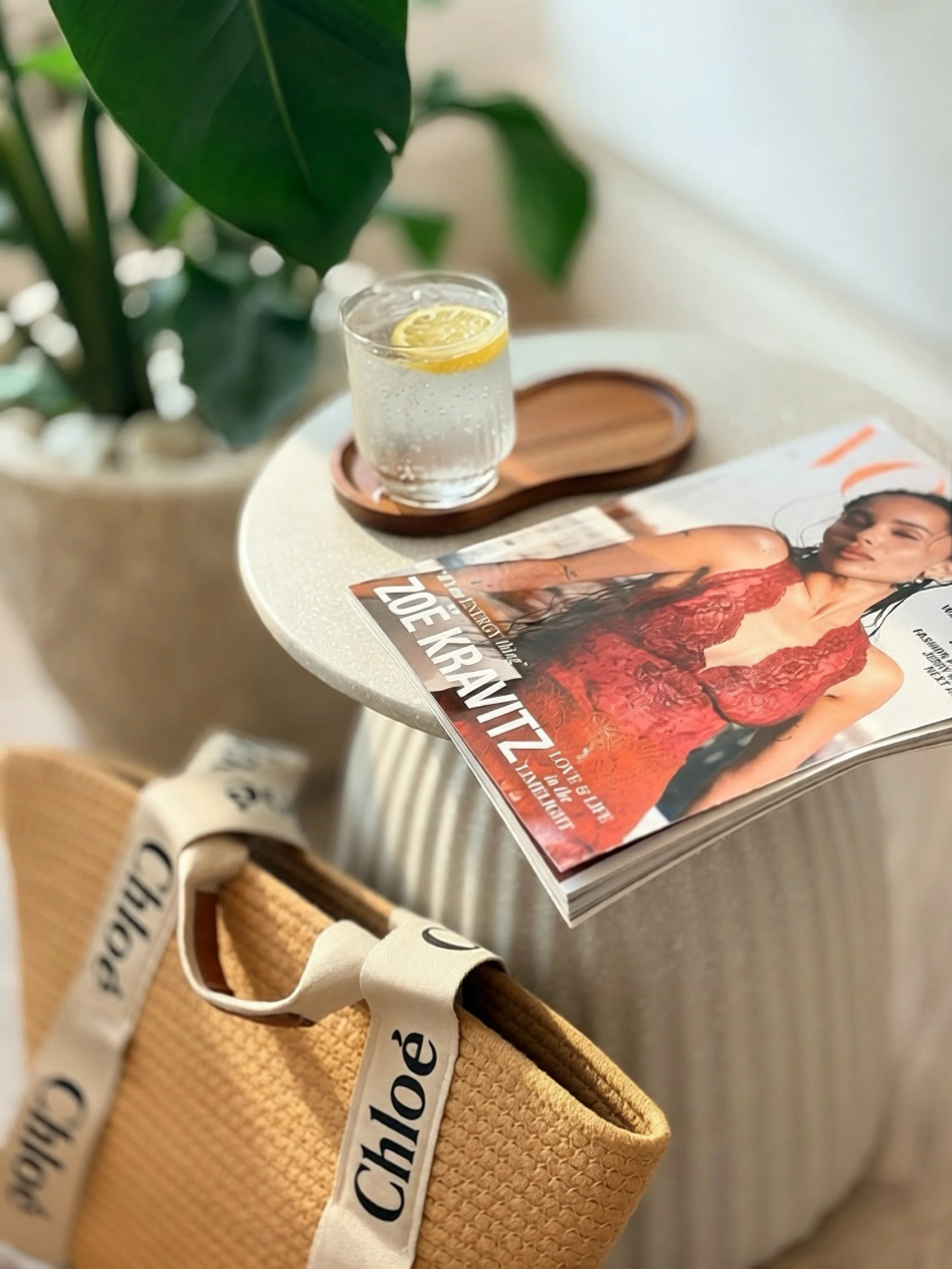 Table with a glass of sparkling water with lemon slice, a fashion magazine, and a tan textured tote bag with black lettering strap.
