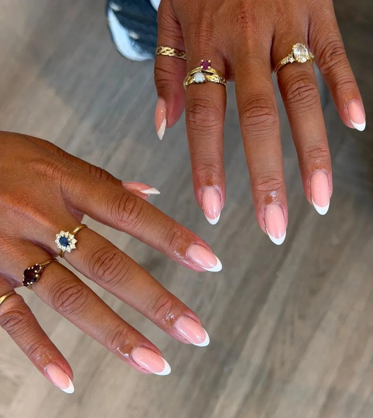 Close-up of two hands with manicured nails and gold rings, some with gemstones, held over a wooden floor.