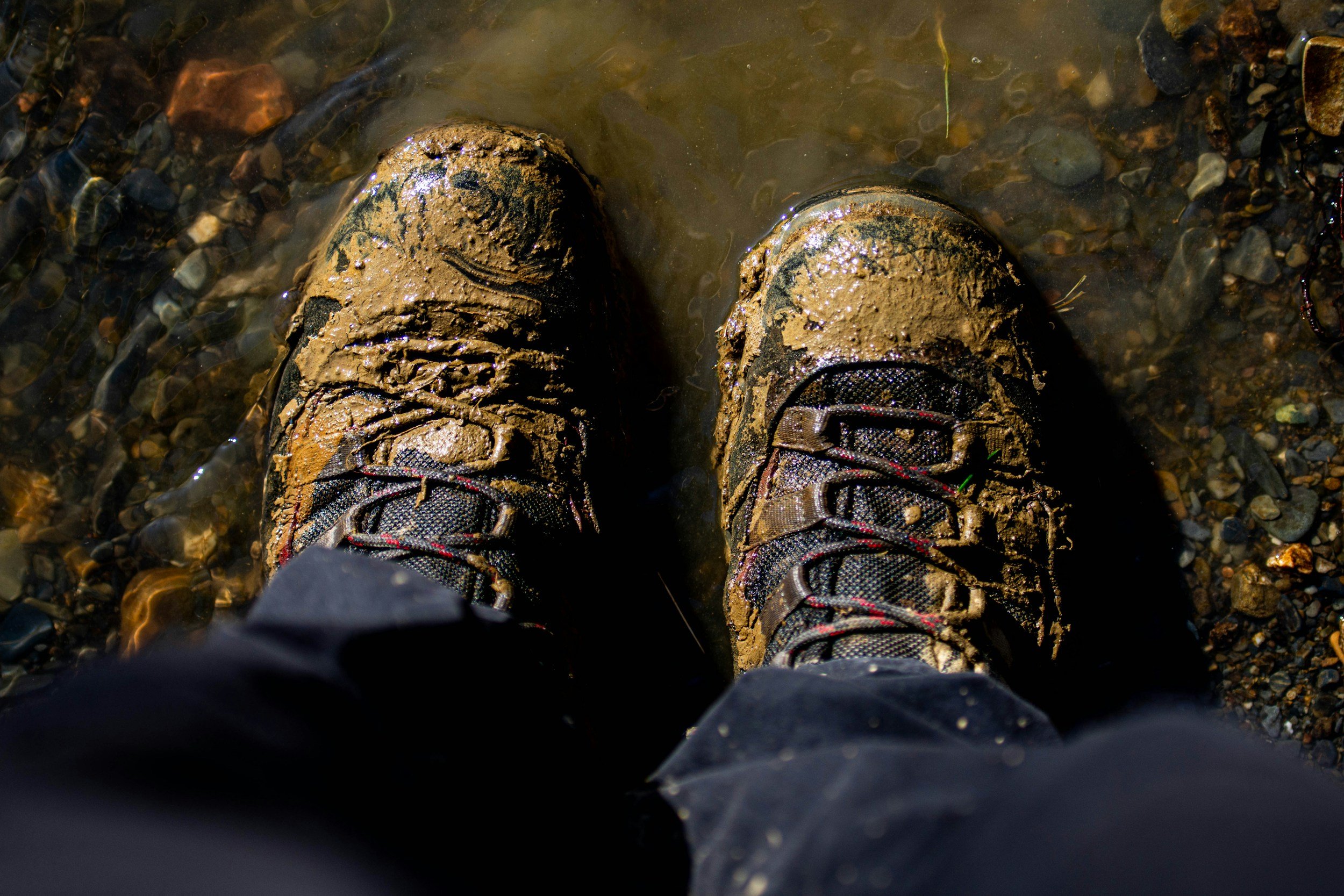 Worn hiking boots covered in mud, standing in shallow water on a rocky surface on a hiking trail near Whistler BC.