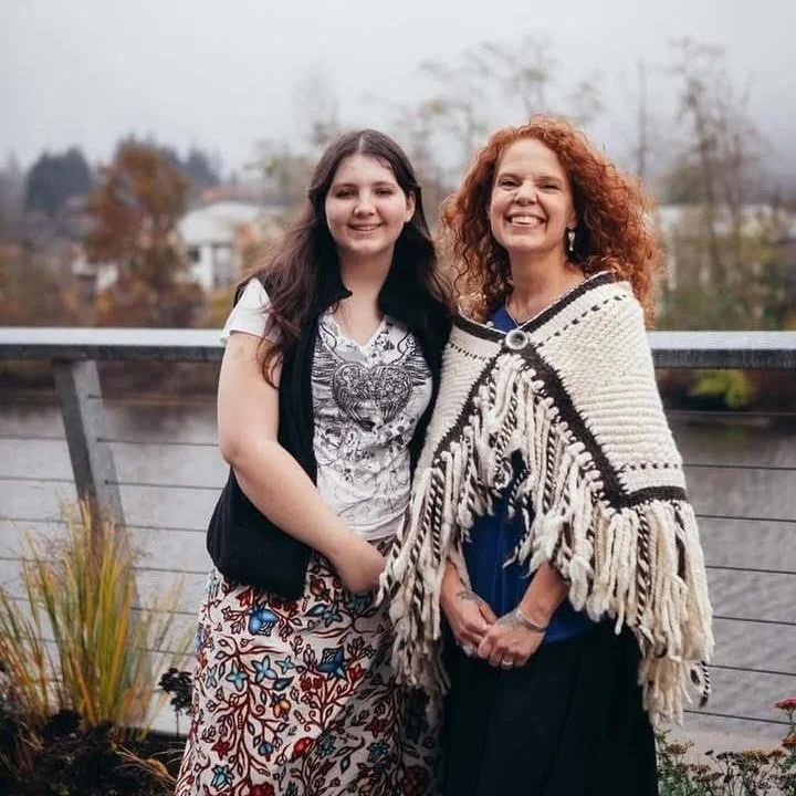 Two women standing outdoors near a body of water, smiling at the camera. One has long dark hair and the other has curly red hair, wearing a fringed shawl.