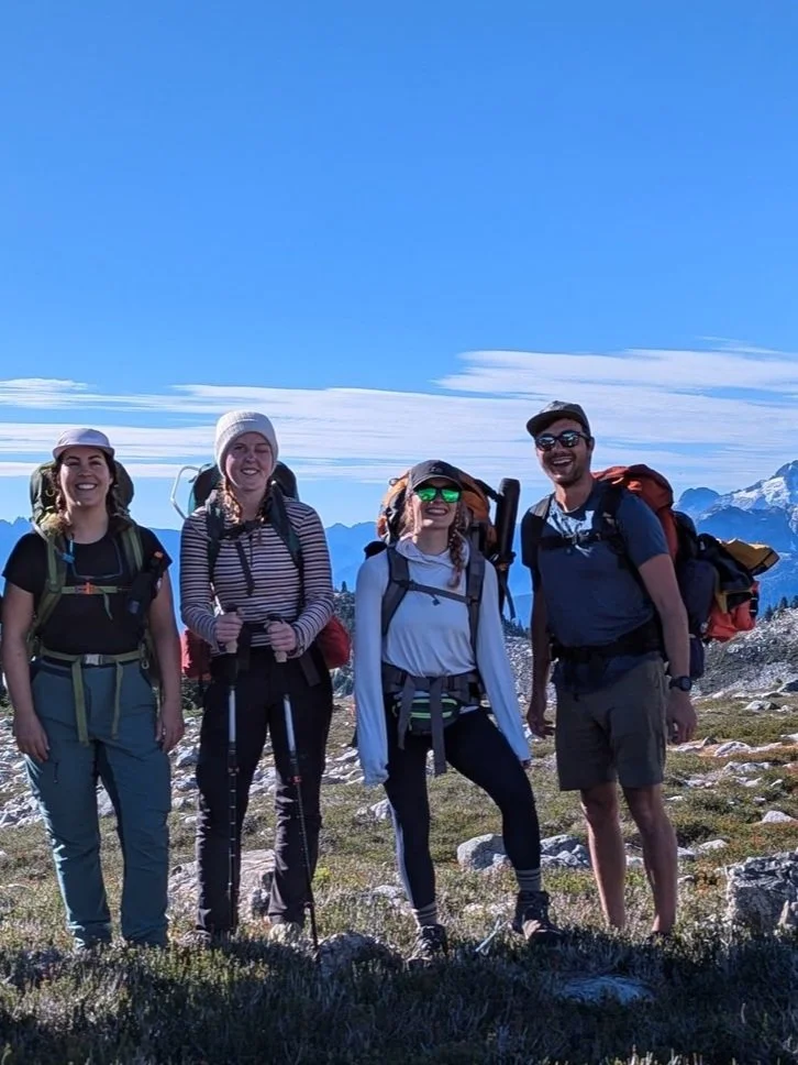 Four hikers standing on a mountain trail with backpacks, smiling, with snow-capped mountains of Whistler in the background under a clear blue sky.