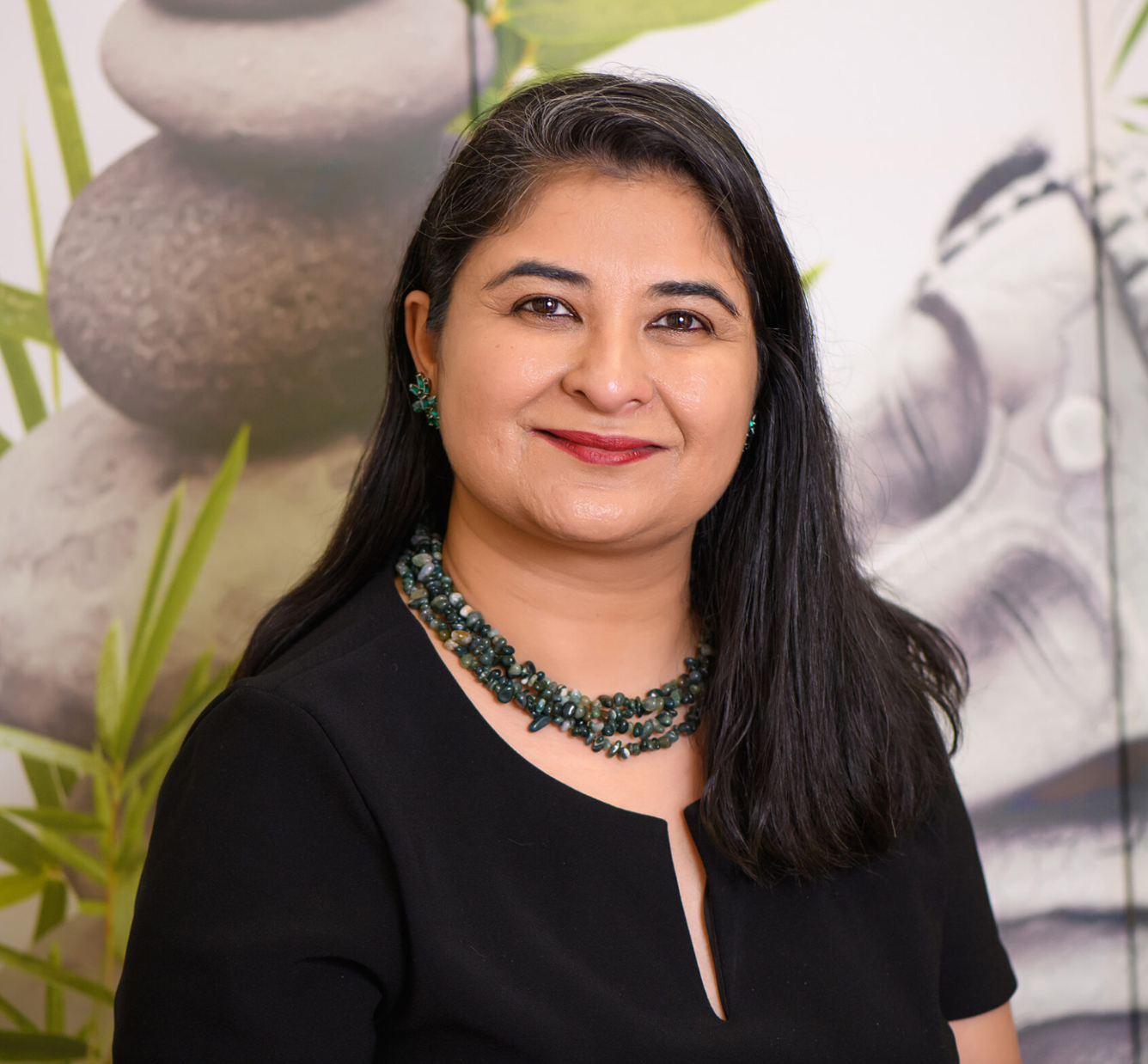 A woman with long dark hair, wearing a black top and green jewelry, smiling for a portrait in front of a decorative background.