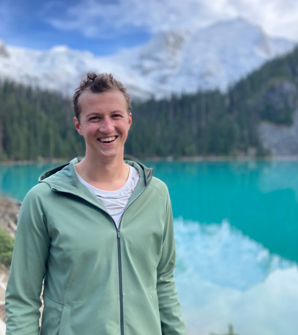 A smiling person standing outdoors near a turquoise lake with snow-capped mountains and evergreen trees in the background.