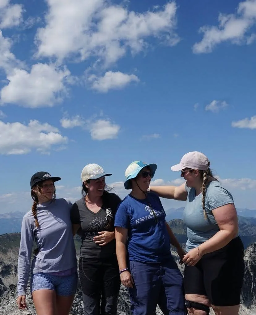 Four women mountain hiking with blue sky and clouds behind them, wearing hats and casual outdoor clothes in the Summer, in Squamish BC.