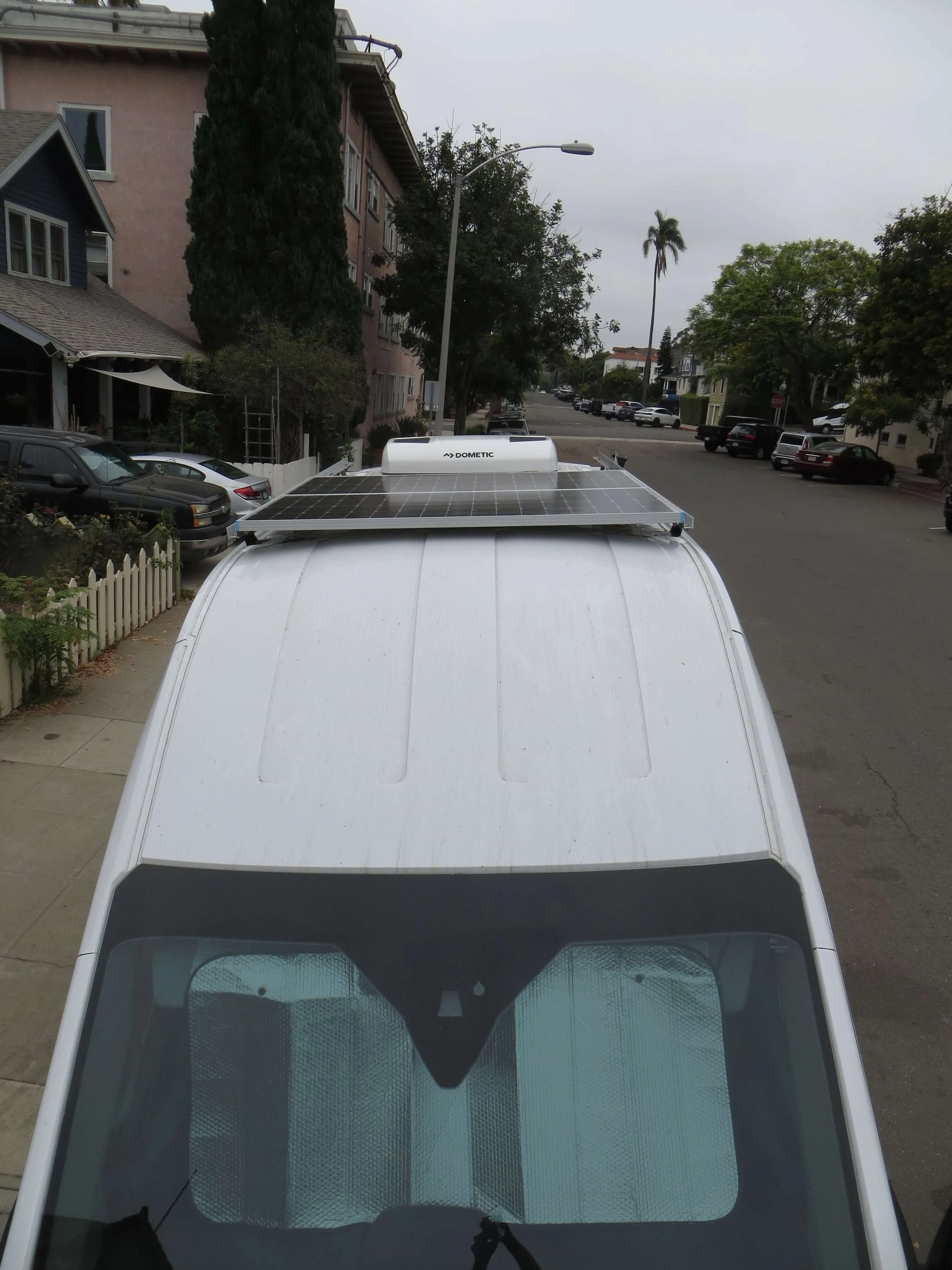 Top view of a white van with a rooftop solar panel and a Dometic air conditioning unit, parked on a residential street with trees and parked cars visible. Example of RV Solar Install near San Diego.  Lithium Battery, DC/DC Converter, LiFePO4, Inverte