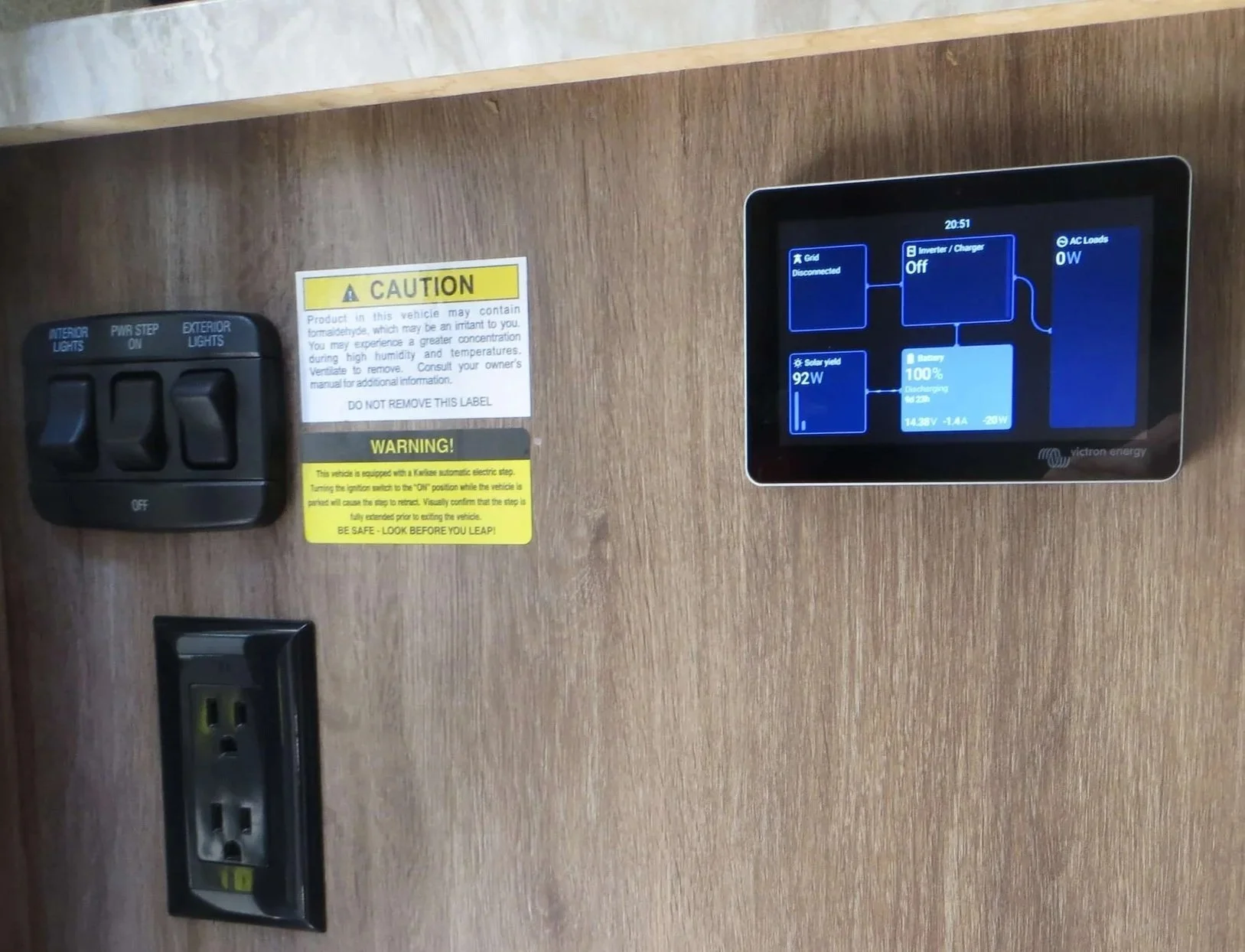 Wooden wall with a black electrical outlet, a switch panel, and a digital energy management display showing solar yield, battery charge, and power status.