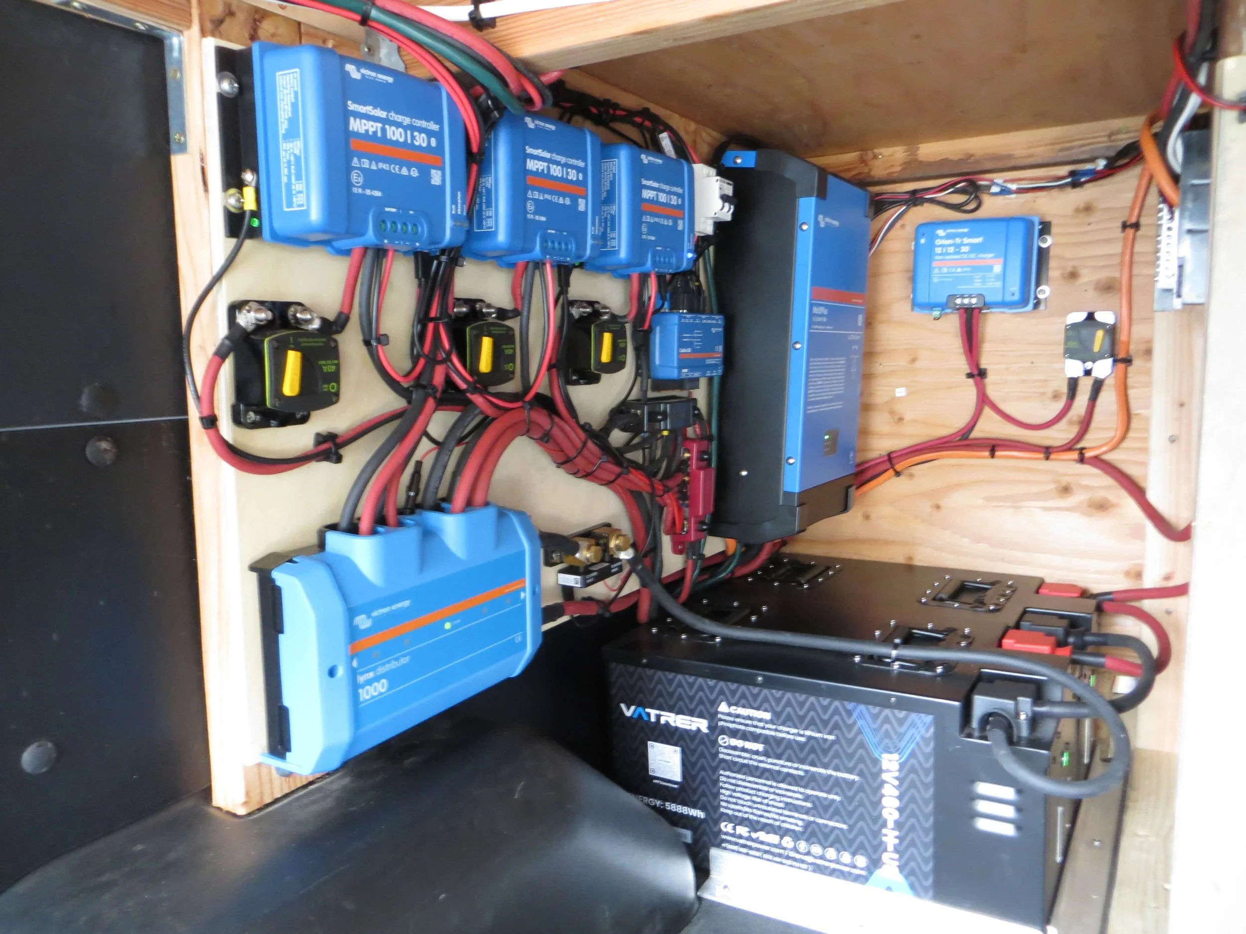 An organized battery and electronic control system inside a wooden enclosure, including blue MPPT charge controllers, a black inverter, a large black battery, Example of RV Solar Install near San Diego.  Lithium Battery, DC/DC Converter, LiFePO4, Inv
