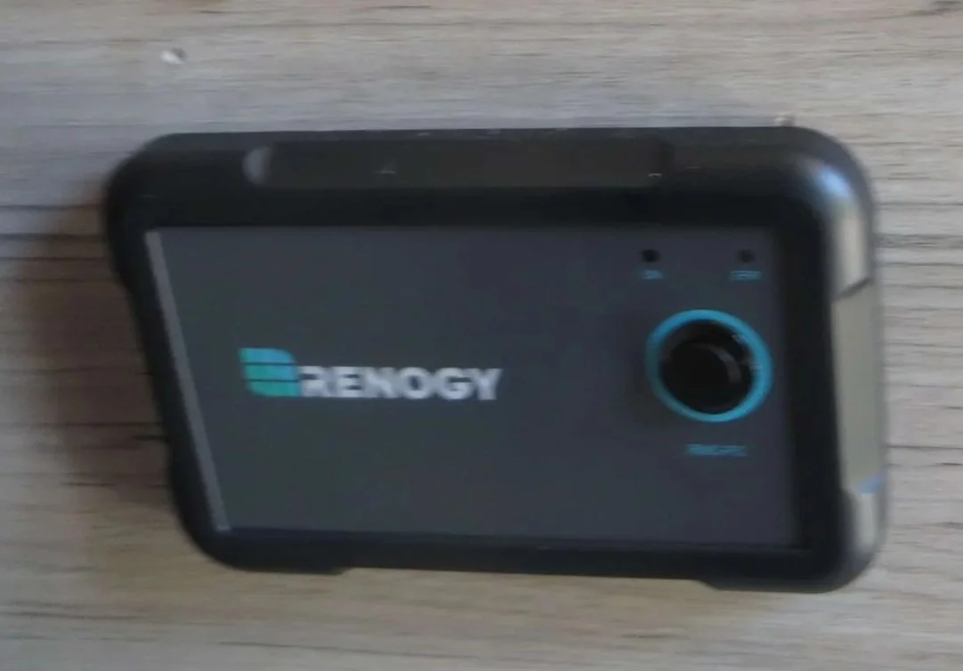 A black rugged mobile phone with a blue circular button and the RENOGY logo on the screen, placed on a wooden surface. Example of RV Solar Install near San Diego.  Lithium Battery, DC/DC Converter, LiFePO4, Inverter, install on RV near San Diego, CA.