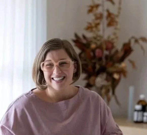 Woman smiling, wearing glasses, sitting indoors with a floral arrangement in the background.