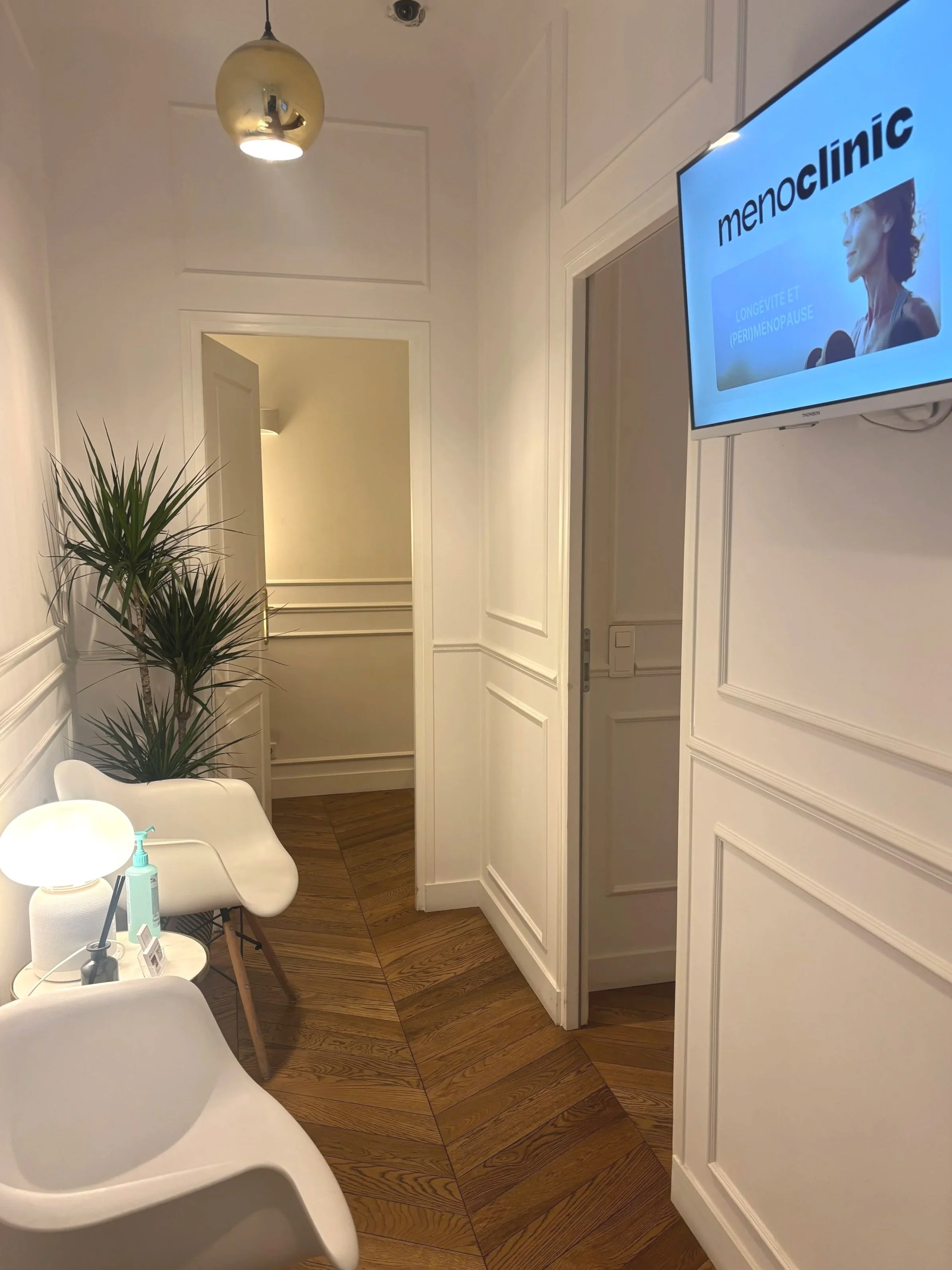 Salle d'attente d'une clinique ou d'un centre de soins avec un fauteuil blanc, une plante verte, une petite lampe de bureau, un distributeur de gel hydroalcoolique, et un écran de télévision affichant menoclinic.