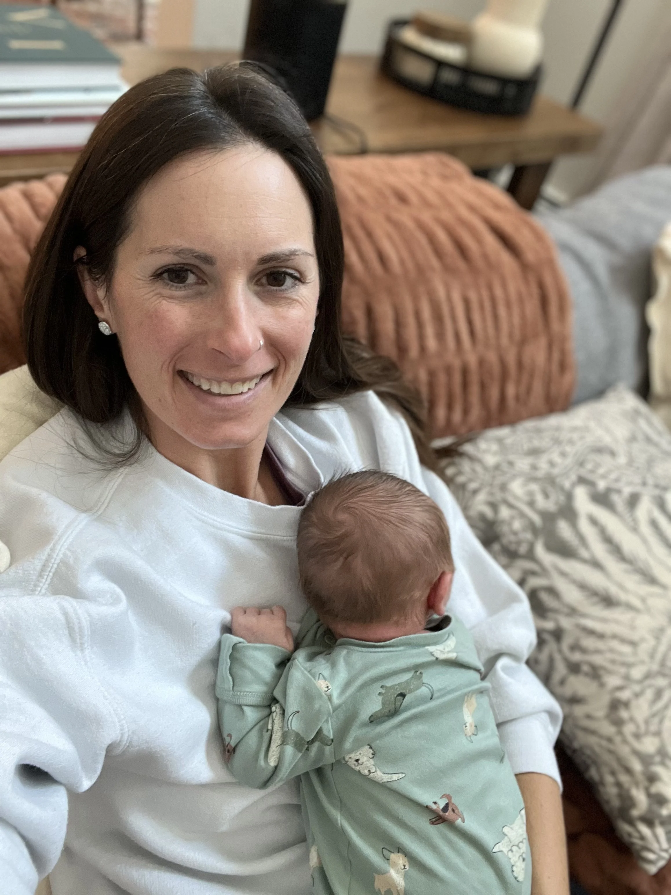 A woman smiling and holding a sleeping baby on her chest in a cozy room with a patterned rug, a plush orange pillow, a wooden table with a stack of books, and some decorative items in the background.