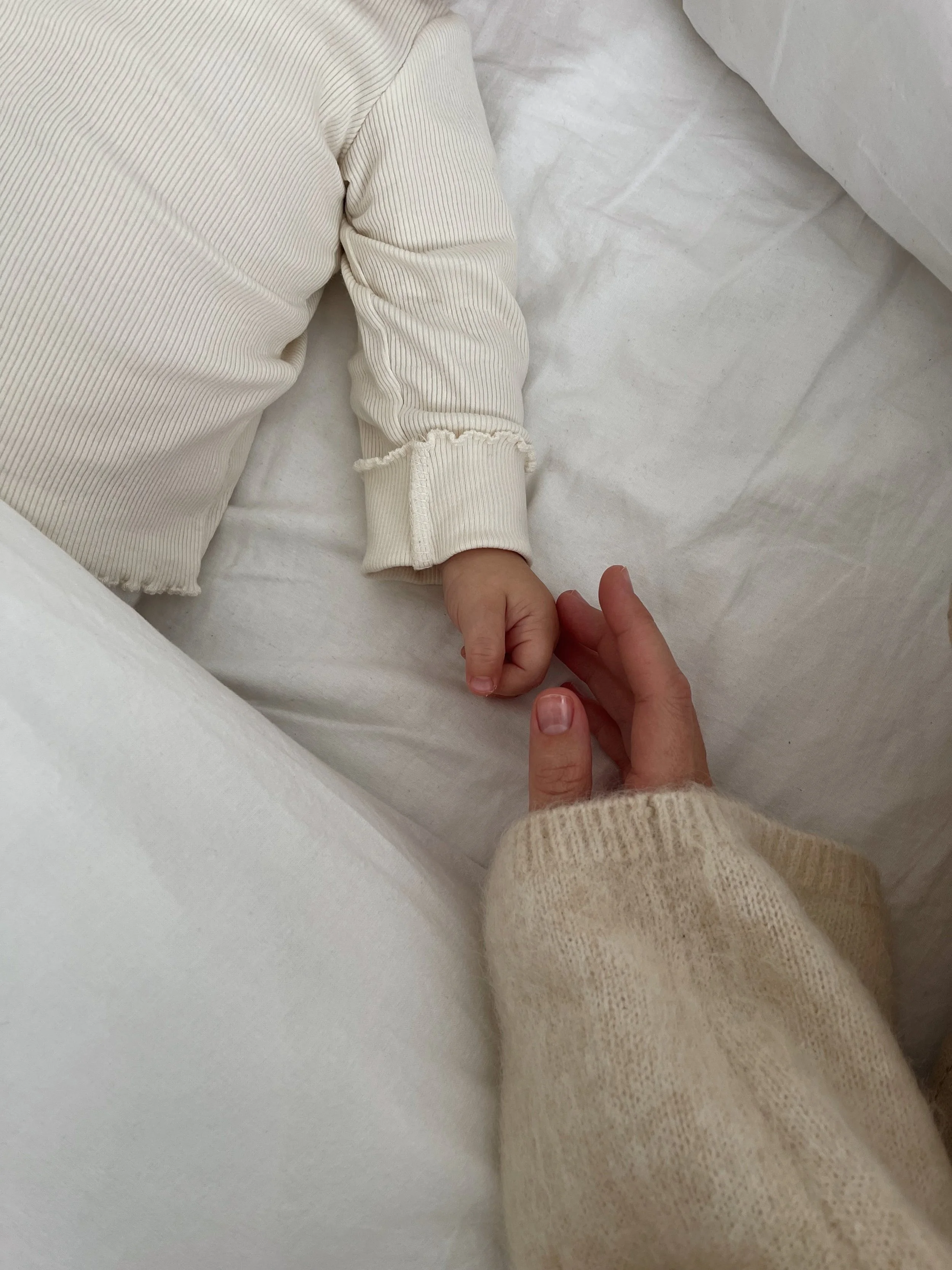 A close-up of a child's hand holding an adult's finger while lying on a white bed with beige and white clothing.