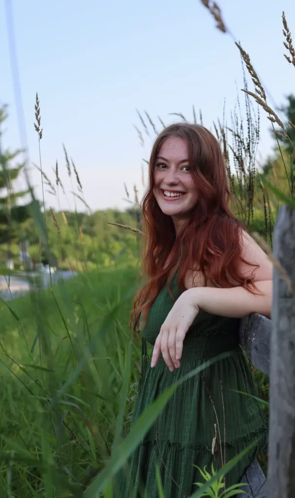 A young woman with long red hair, smiling and leaning on a wooden post in a grassy outdoor setting with trees in the background.