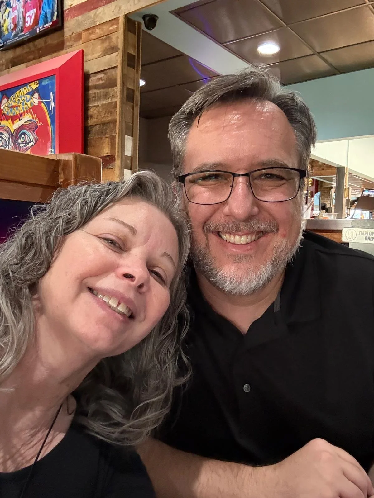 A smiling man with glasses and a woman with gray hair taking a selfie in a restaurant or bar with wooden walls and colorful artwork in the background.