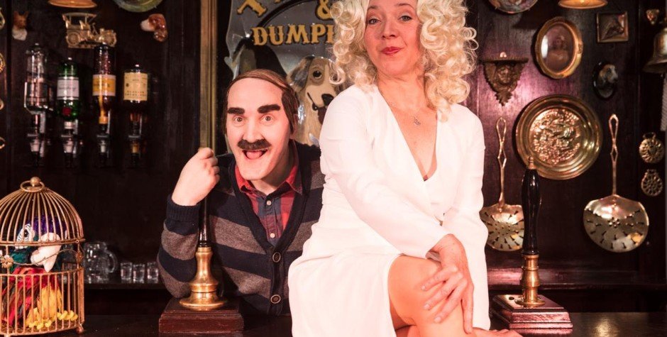  Old man landlord character with bushy eyebrows and moustache with a woman sitting on the bar in white 