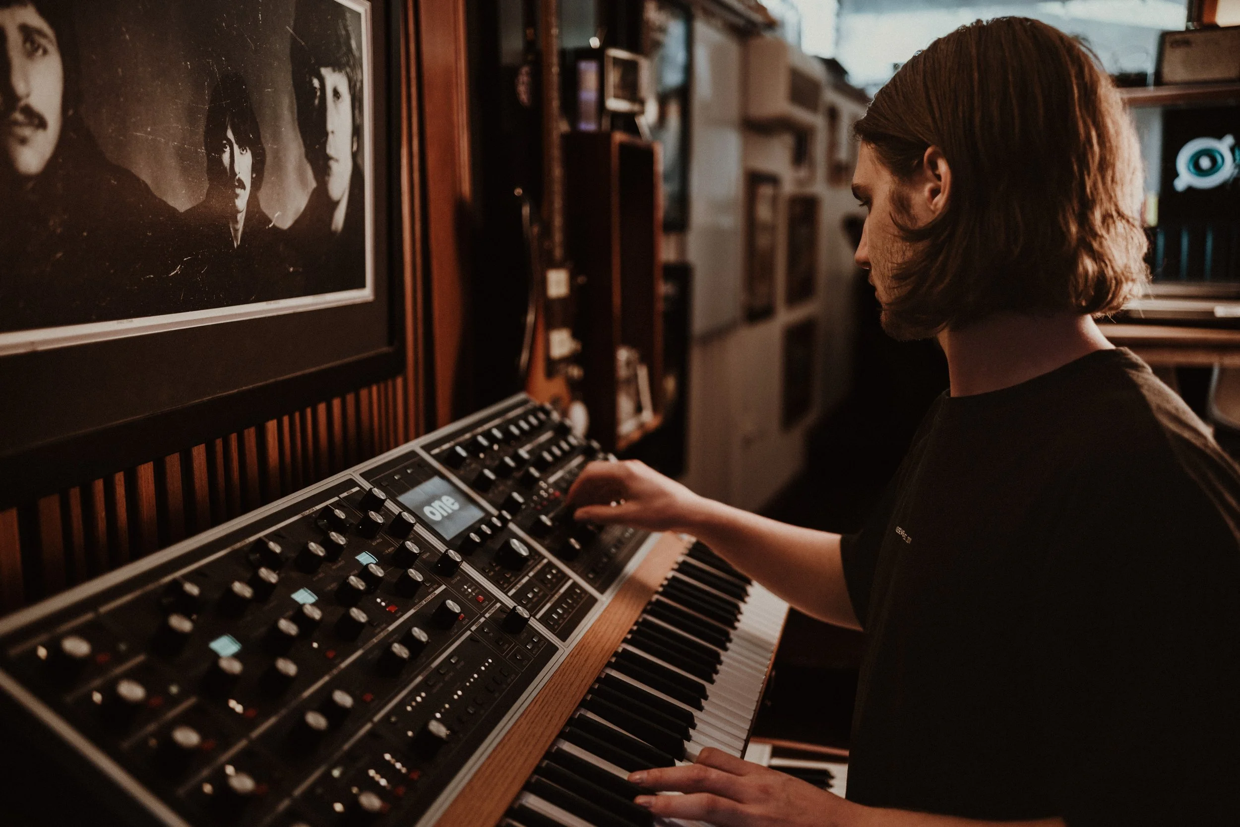 A person with shoulder-length hair playing a vintage electronic keyboard in a room decorated with musical posters and framed photographs.