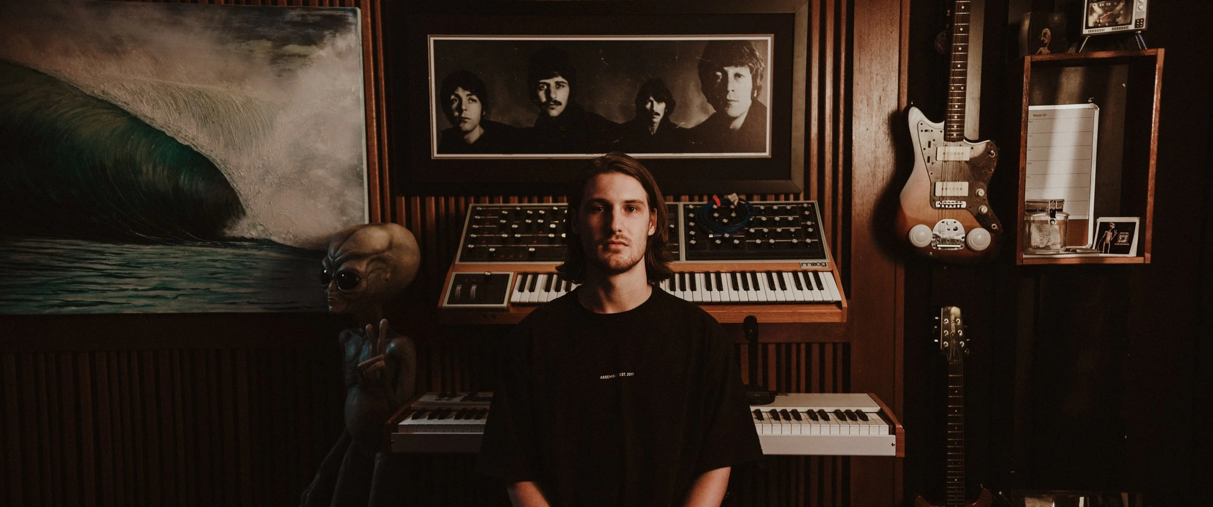Young man sitting in a music studio with guitars, artwork, and a framed photo of The Beatles in the background.