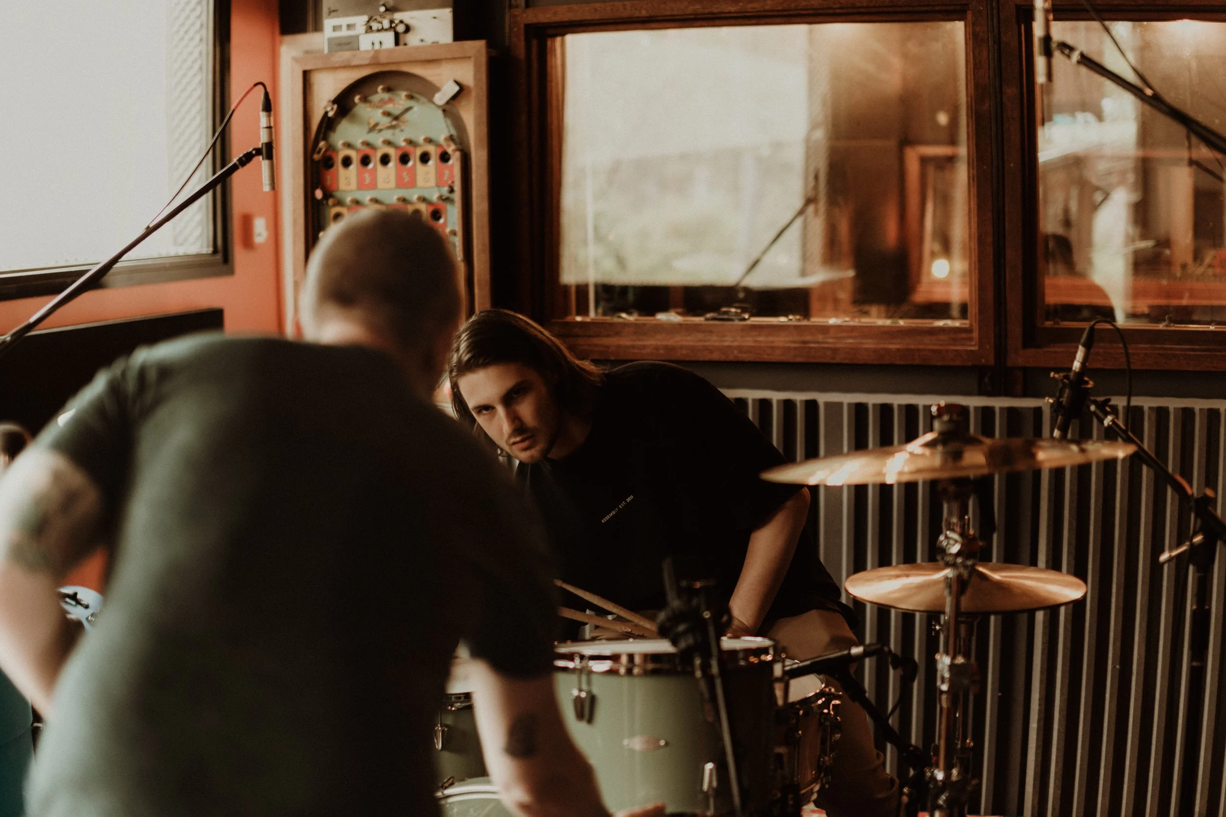 Two men in a music studio, one sitting behind a drum set and the other leaning forward, with recording equipment and acoustic panels visible in the background.