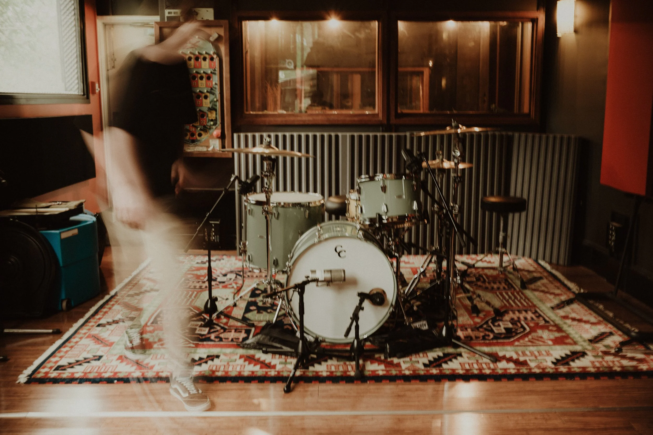 A music recording studio with a drum set on a patterned rug, surrounded by microphones, a stool, and recording equipment, with a person moving nearby and a blurred background.