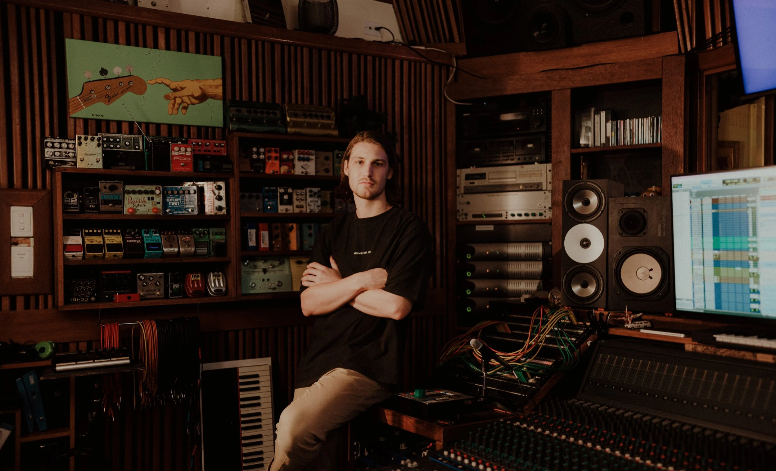Young man standing with arms crossed in music production studio with audio equipment, mixing console, speakers, and a computer screen displaying a digital audio workstation.